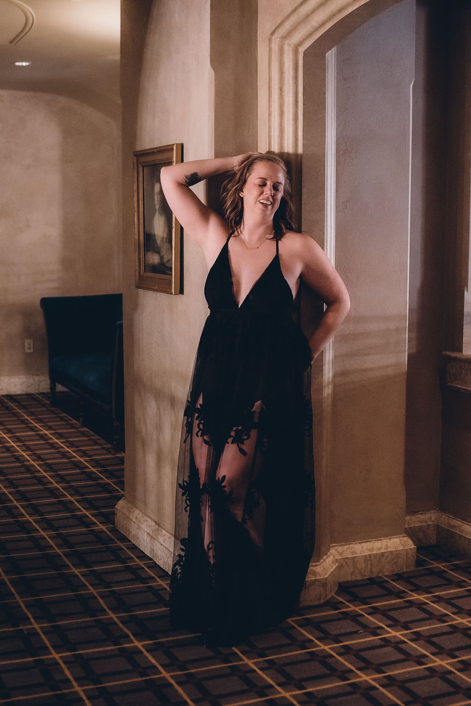 Woman in black sheer lace gown posing elegantly in a warmly lit hotel corridor with ornate decor.