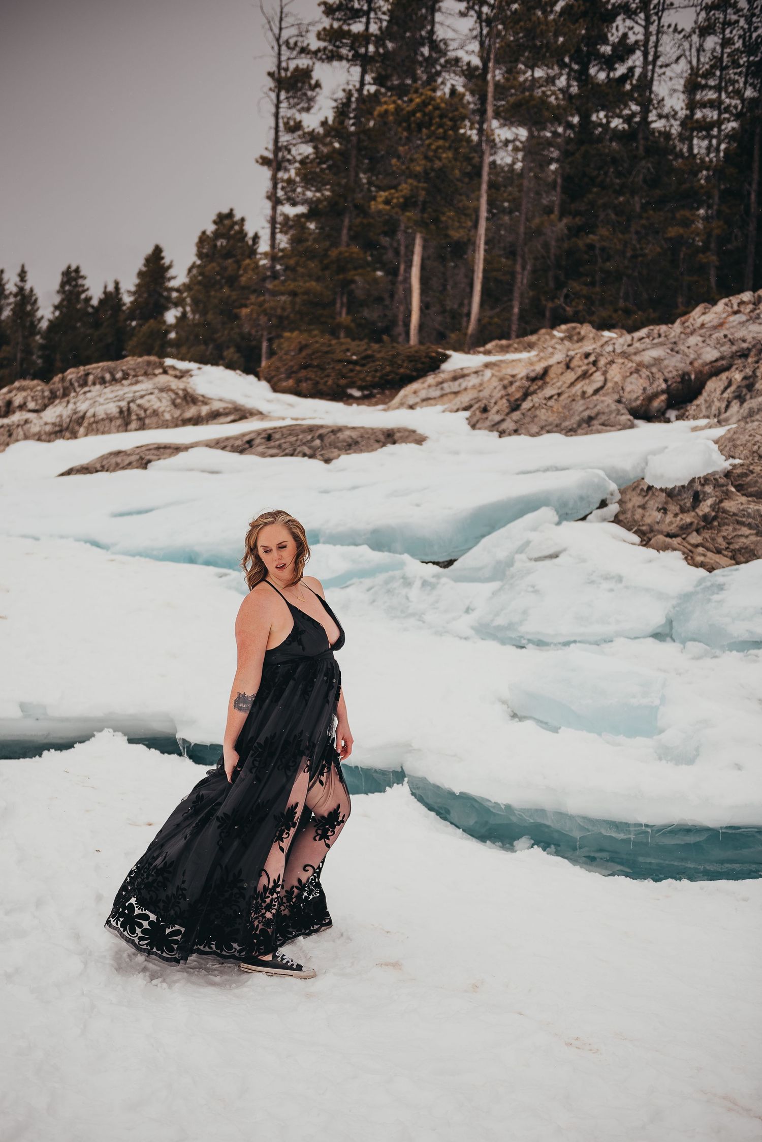 Woman in black lace gown poses among blue ice formations and snow with pine trees in background.