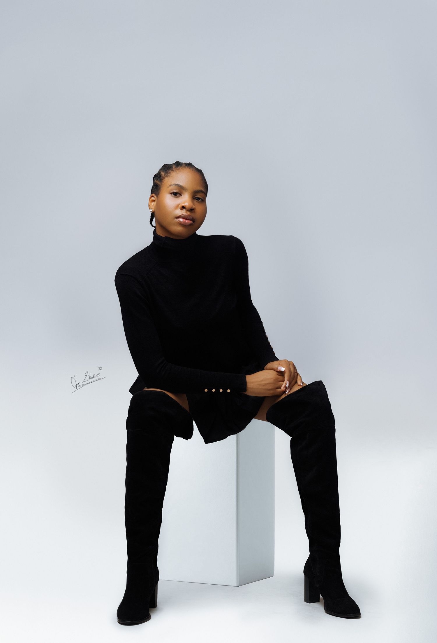 A portrait of a beautiful Nigerian model sitting on the studio box in a black turtle neck, black skirt and black knee high boots