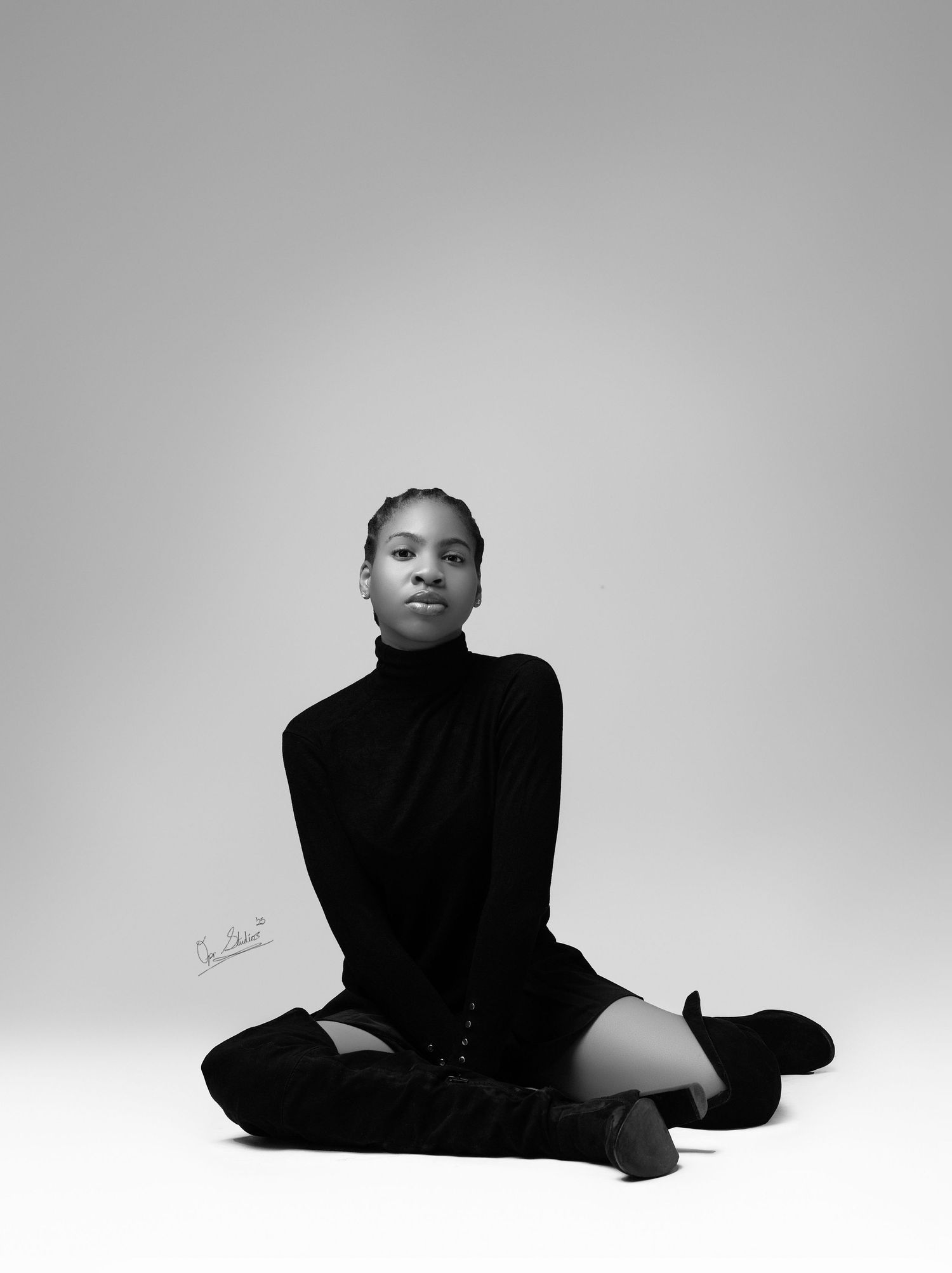 A B & W portrait of a beautiful Nigerian model sitting on the studio floor in a black turtle neck, black skirt & black knee high boots