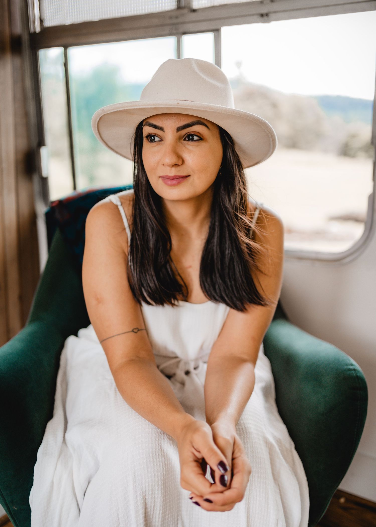 Mari Diwo, Studio Diwo photographer, sitting indoors in a white outfit and beige hat, natural light portrait.