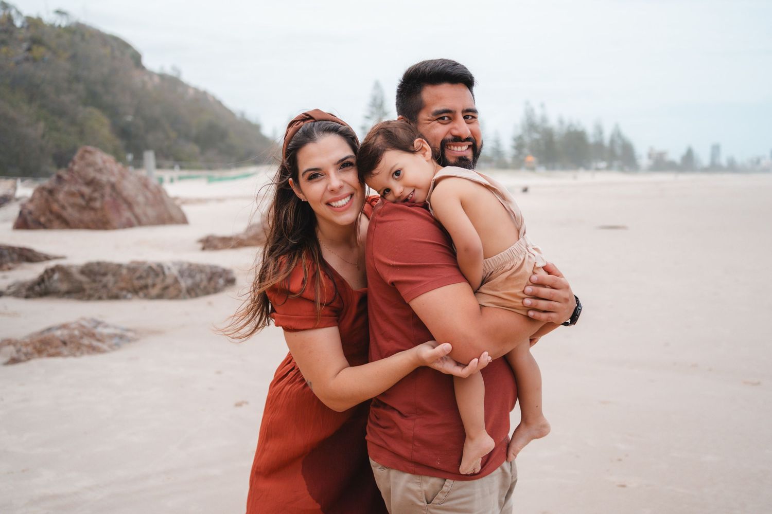 Pamela, Marcos and Otto enjoying a beach family session, warm hugs and natural smiles with ocean and coastline in the background.