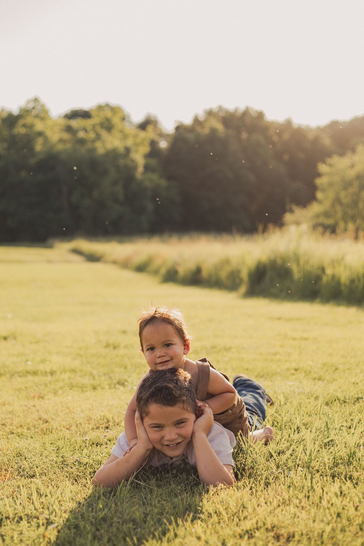 Two young boys in a bright, open field in the summertime, the youngest playfully on top of his brother laughing and smiling.