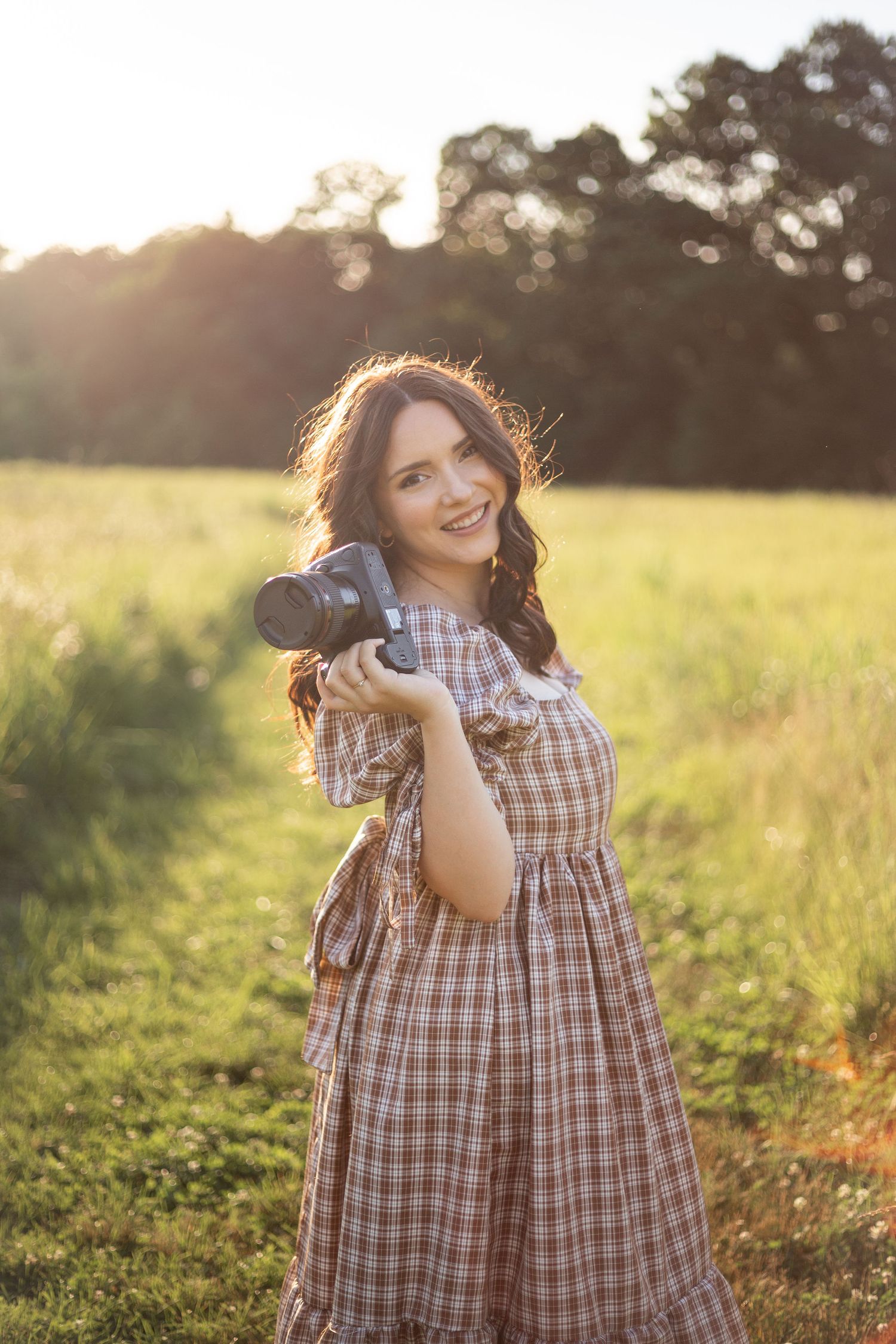 A photographer smiling at the camera and holding her camera in her hand in an open sunny field.