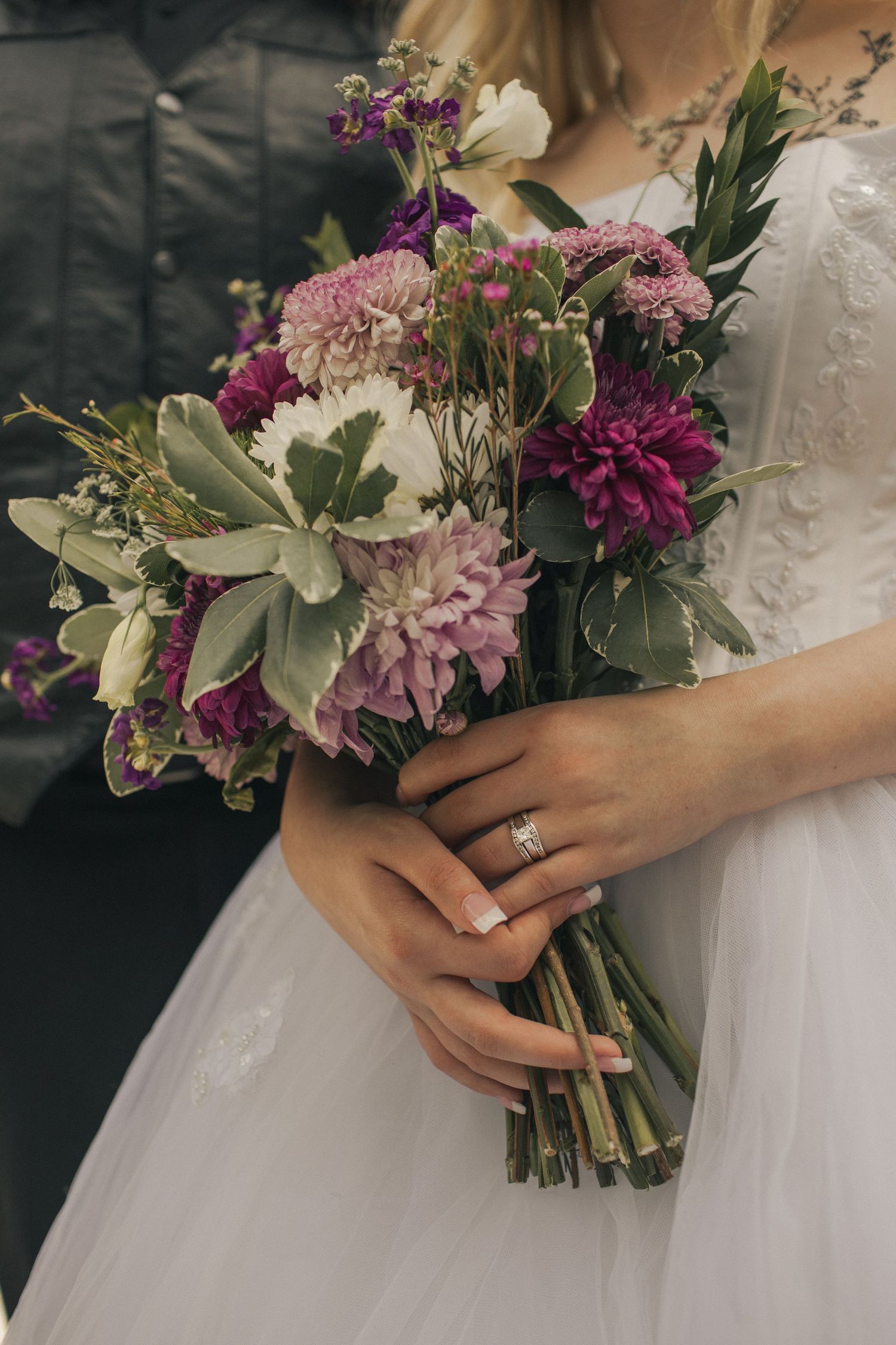 A close-up image of a bride's bouquet, with the groom standing next to her; her wedding ring visible as she holds the flowers.