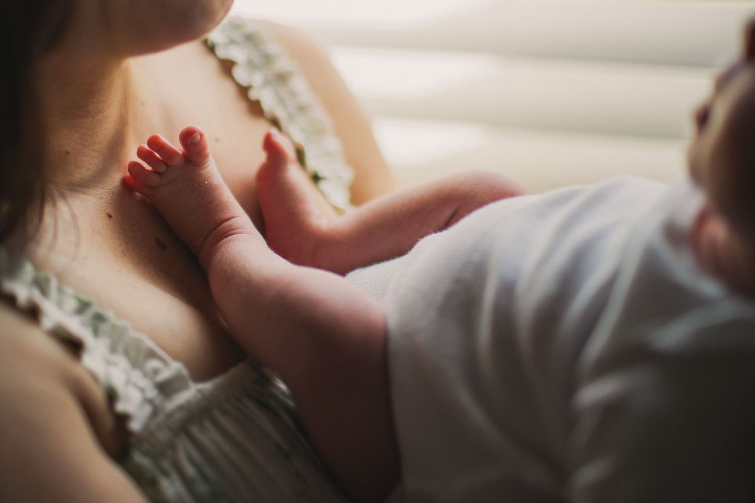 A closeup of a newborn baby's feet on his mother's chest in front of a window in a lifestyle newborn session.