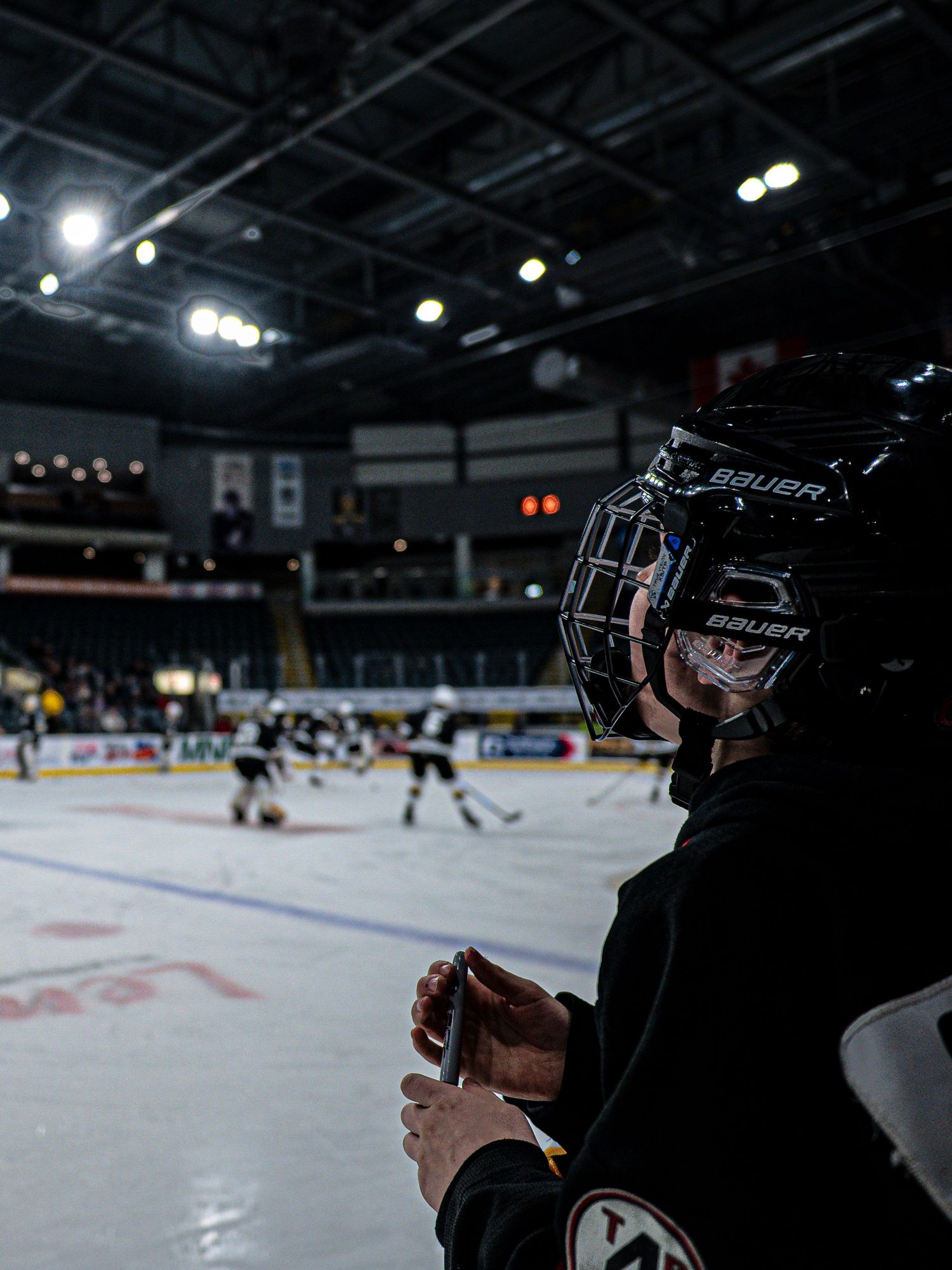 Hockey player watches game from bench in darkened indoor arena.