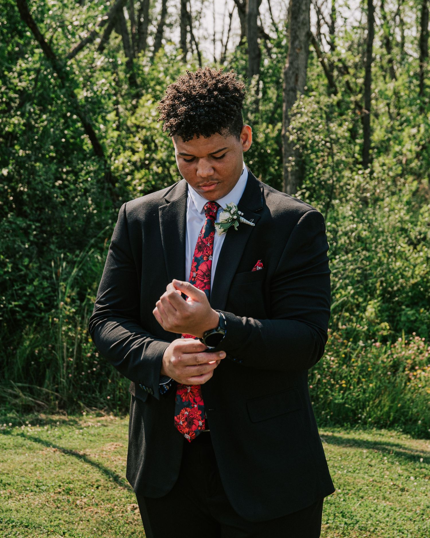 A person in a black suit and red tie checks their phone while standing outdoors in a wooded area.