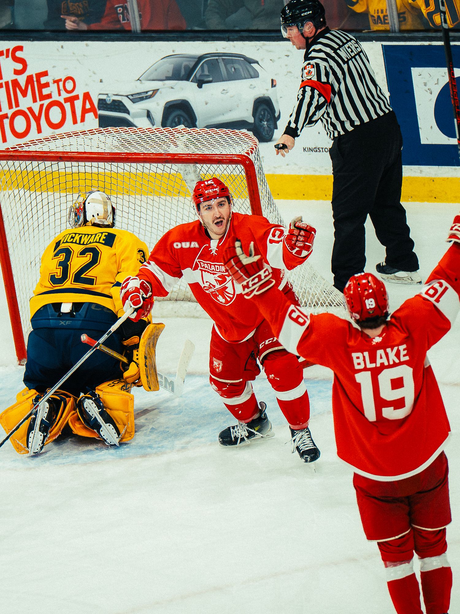 Hockey players in red jerseys celebrate near the goal while facing a goalie in yellow jersey number 32 during a game.