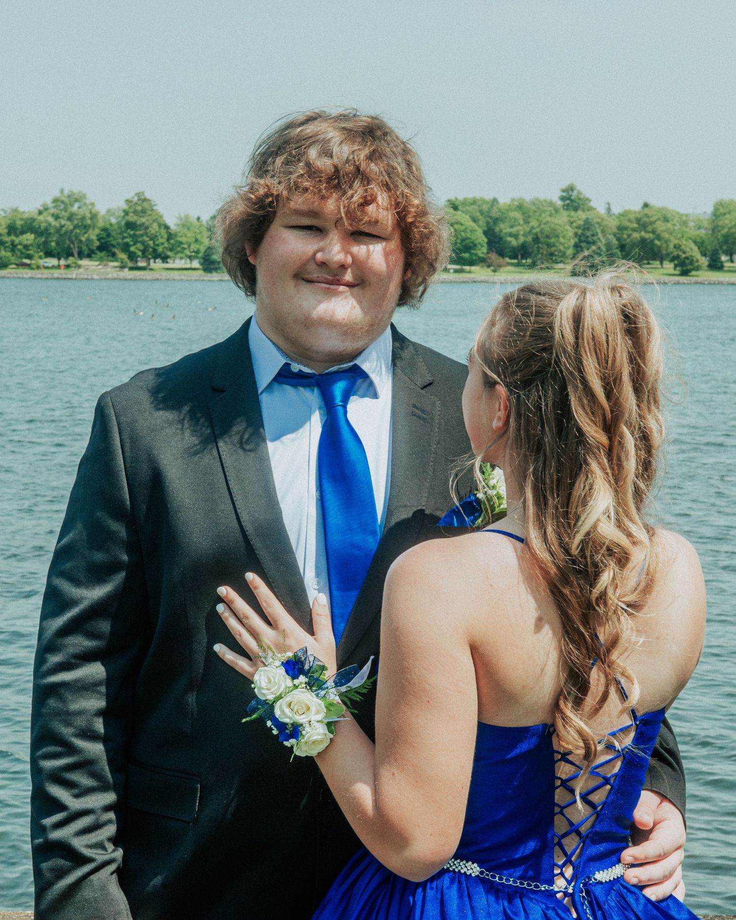 A couple in formal attire poses together at the waterfront, with the person in blue showing off a ring.