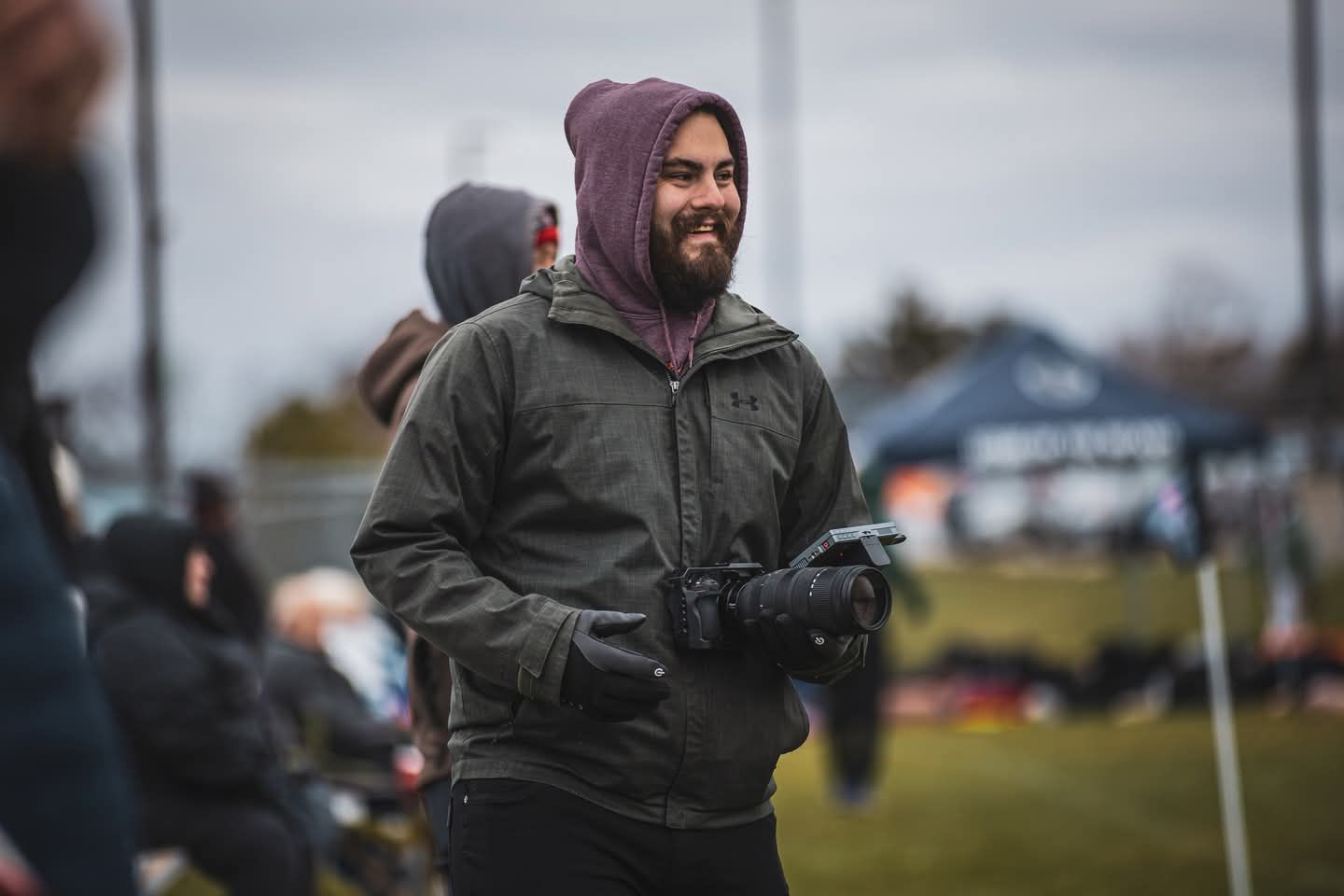 Smiling bearded man in green jacket and purple hoodie wearing black gloves outdoors at a sports field.