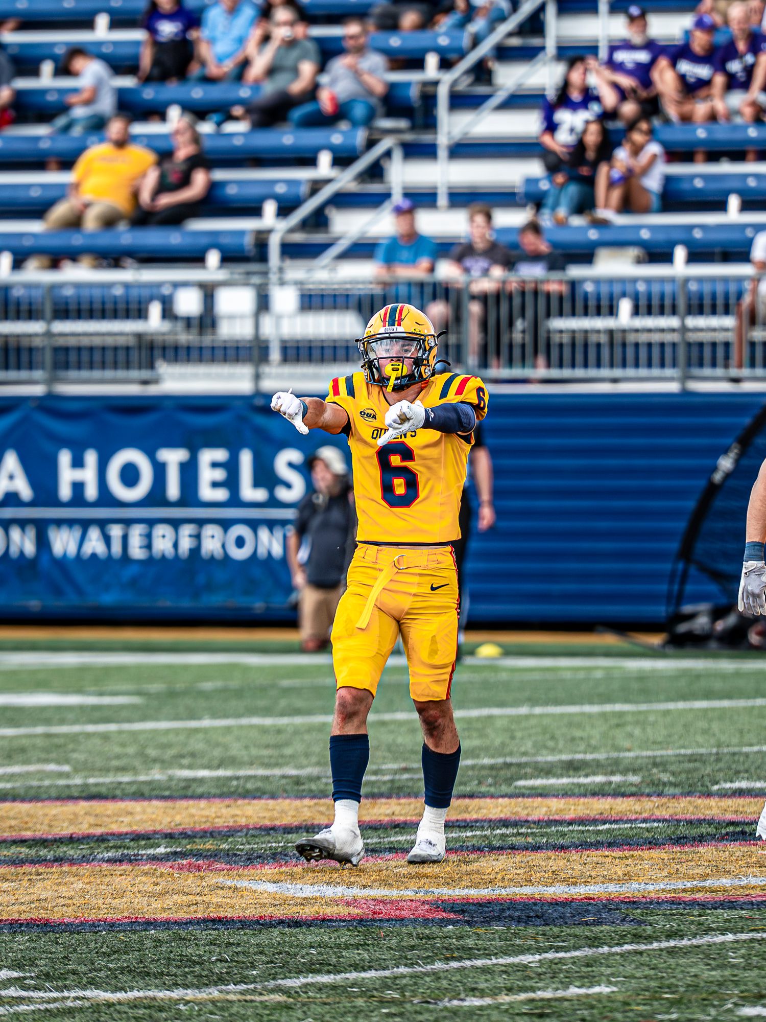 Football player in yellow uniform stands in ready position on field during game against blue backdrop.