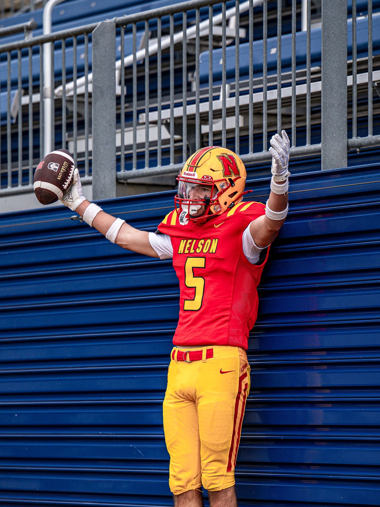 Football player in red and yellow uniform raises arms while standing in front of blue stadium bleachers.