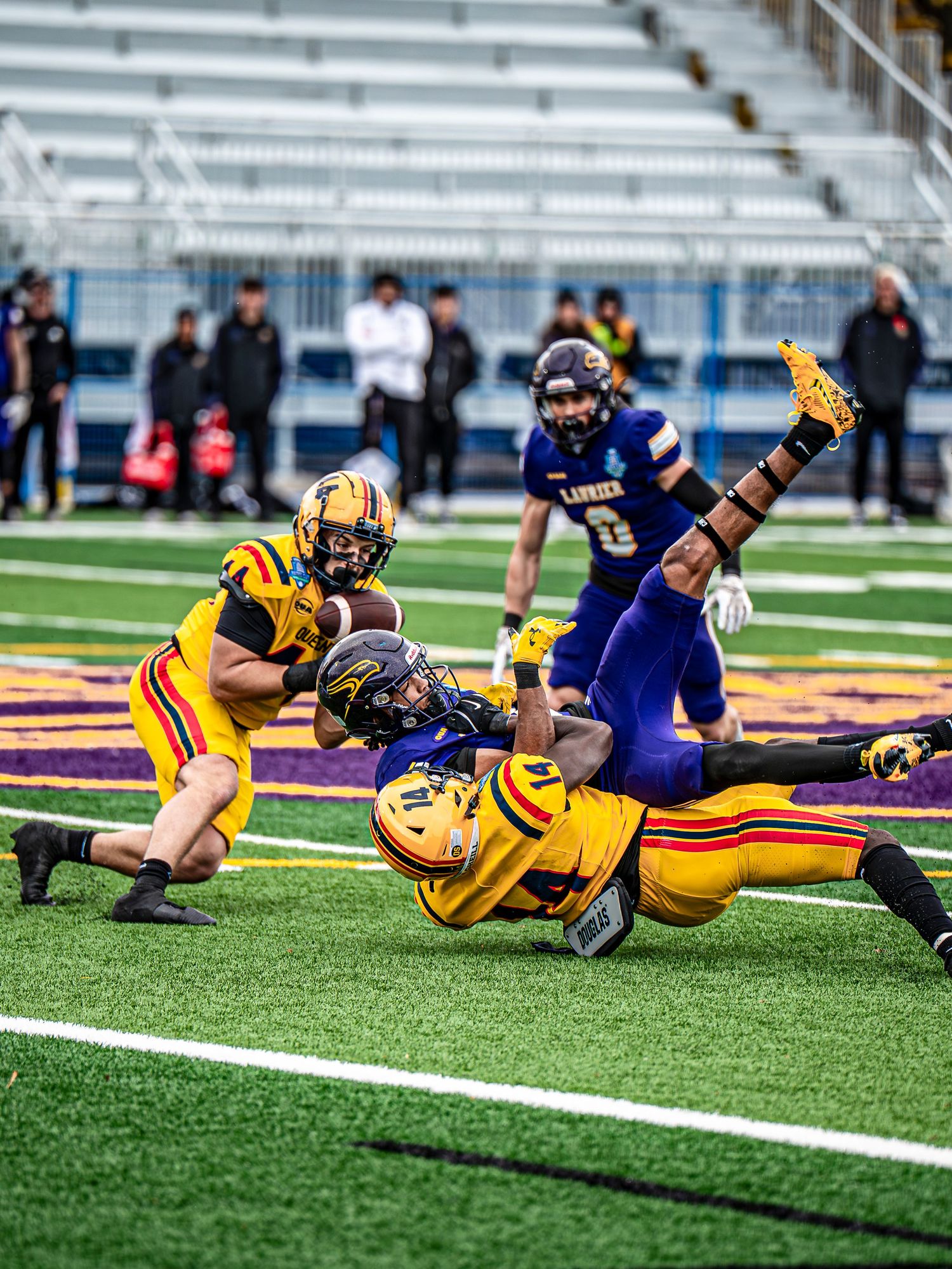 Players in yellow and blue uniforms dive for tackle during football game on artificial turf.