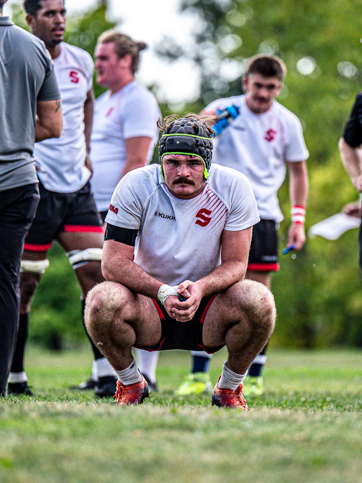 Rugby players crouch on the sideline of a grass field during practice.