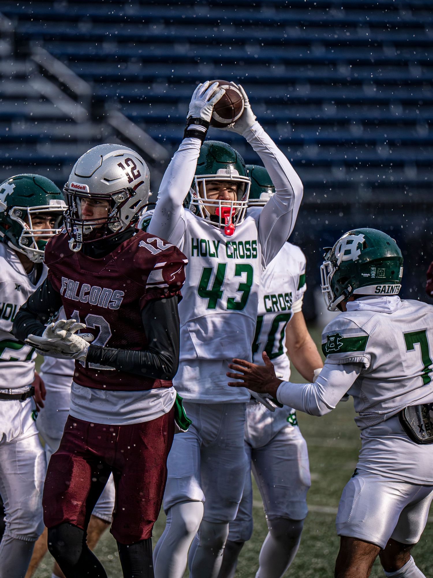 Football players from opposing teams in maroon and white uniforms compete during a snowy game.