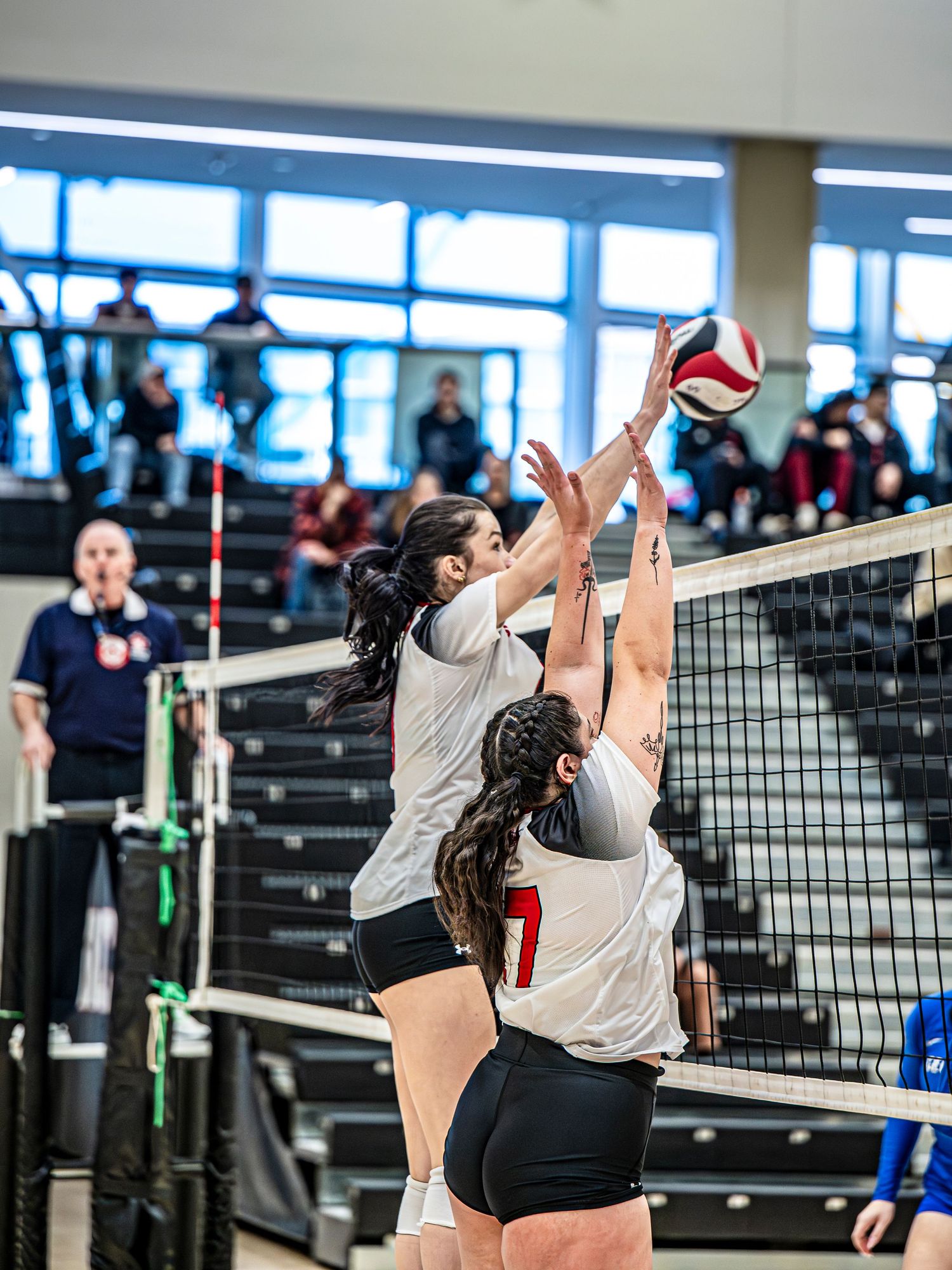 Volleyball players leap high to block a shot at the net during an indoor match.
