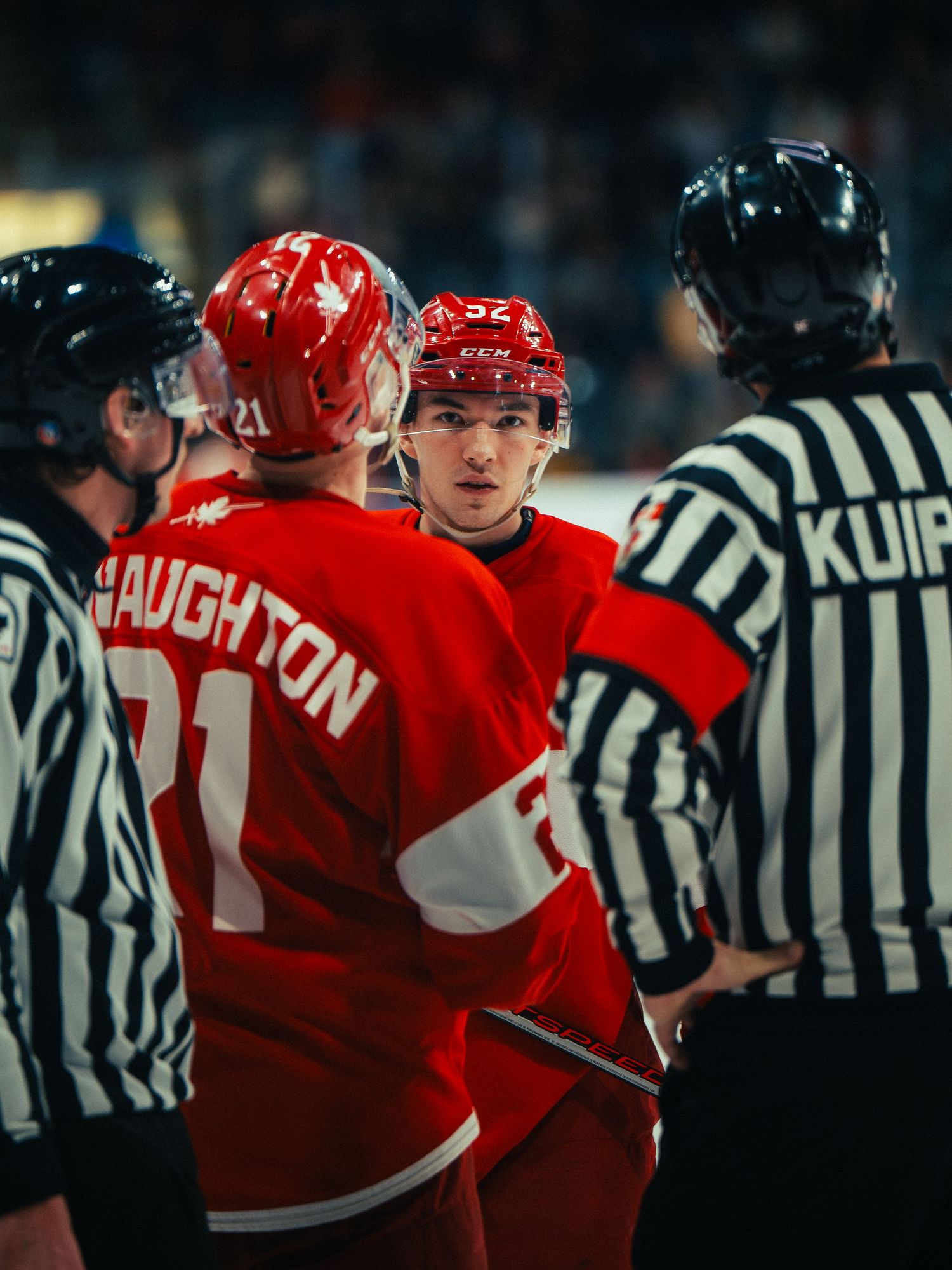 Hockey players in red jerseys gather around referees in black and white striped uniforms during an on-ice discussion.