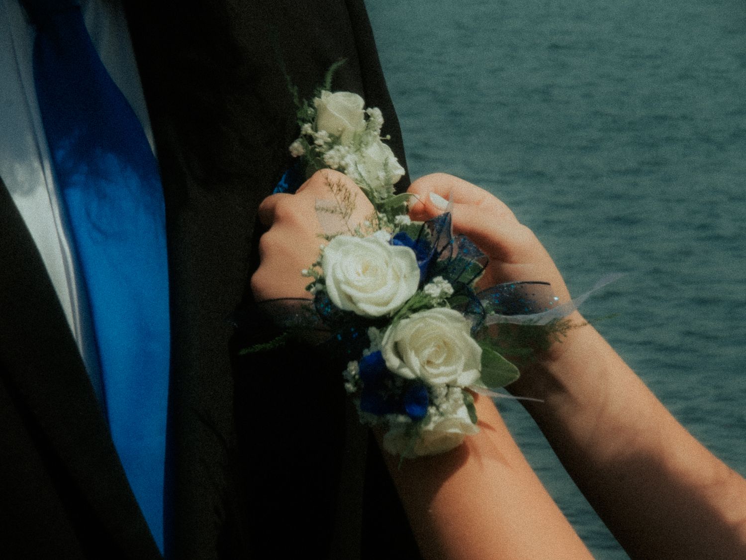 Close-up of hands pinning a white rose corsage with blue ribbon against a teal water background.