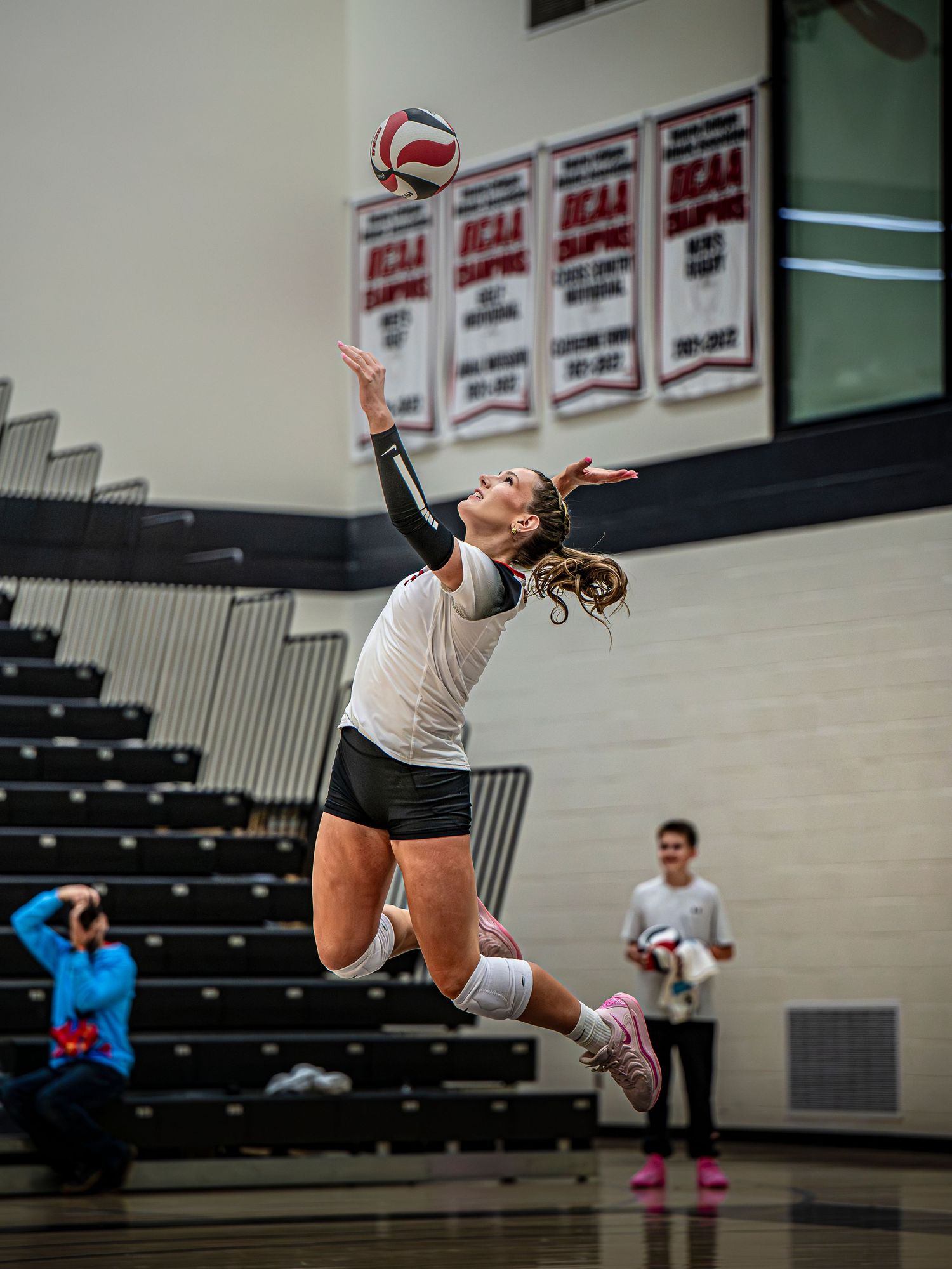 Volleyball player leaps to serve ball in indoor gymnasium with championship banners.