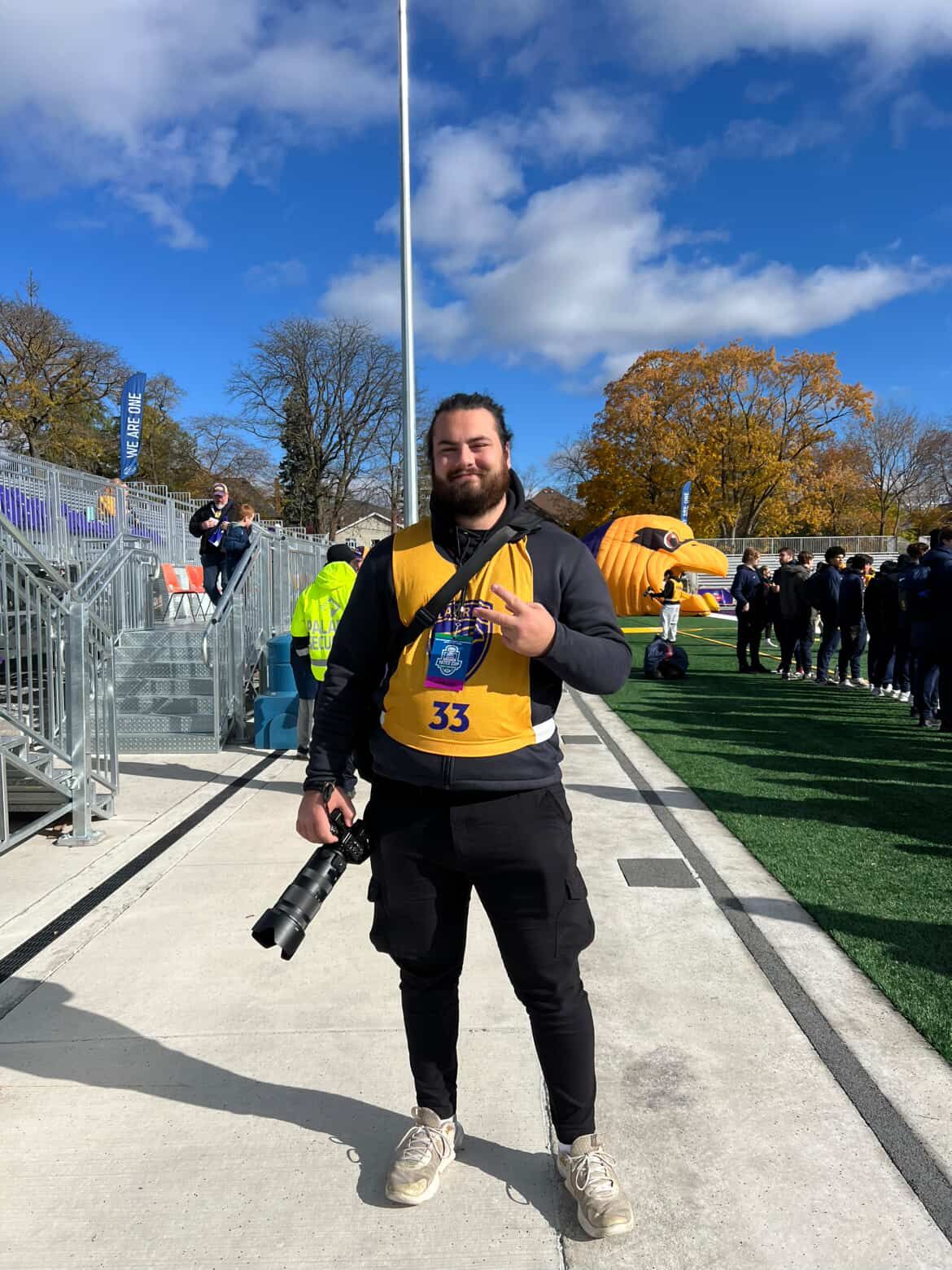 A man in a yellow and black jersey number 33 holds a camera at a football field with a mascot behind him.