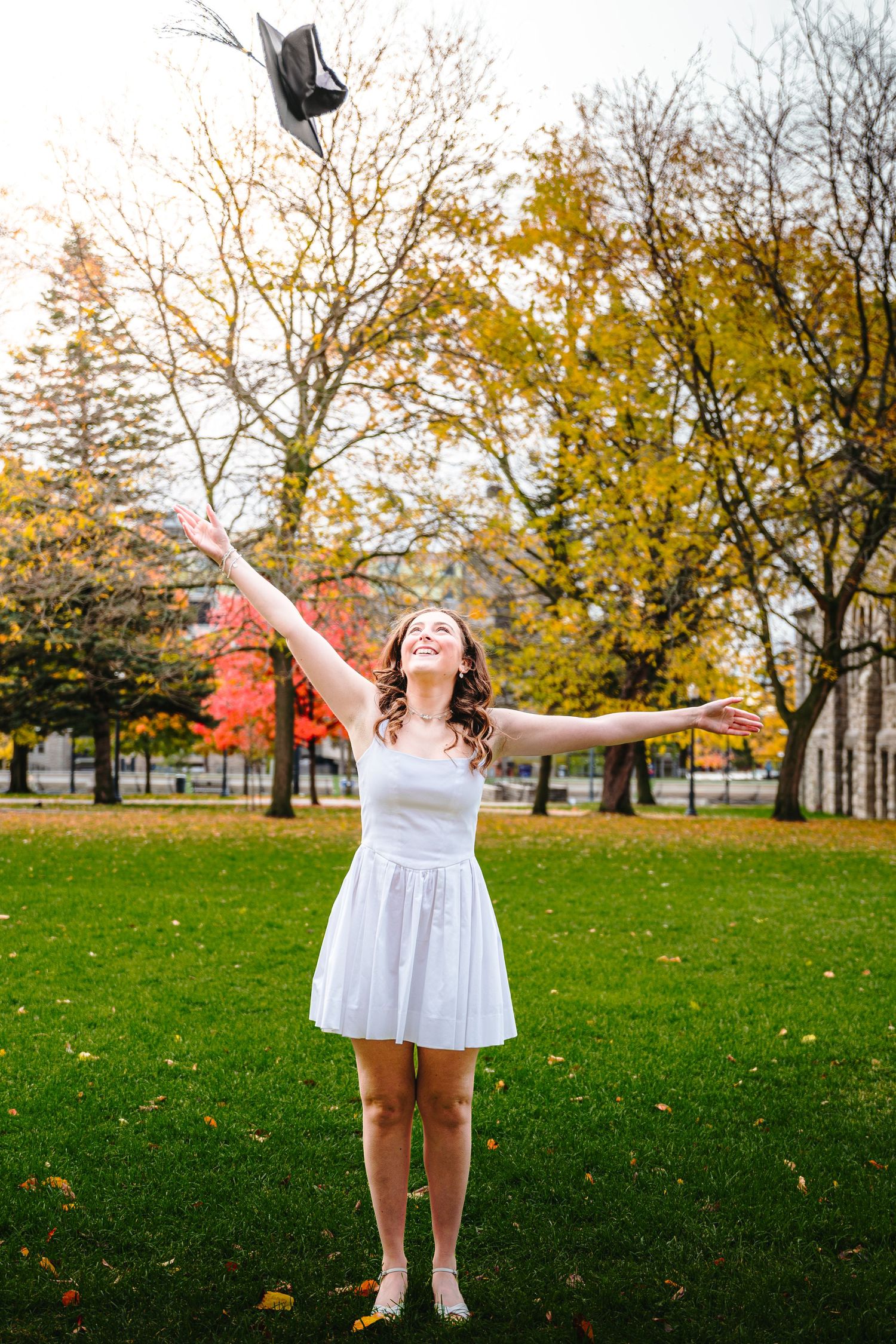 A joyful figure in white dress poses with outstretched arms in a park with autumn trees and fallen leaves.