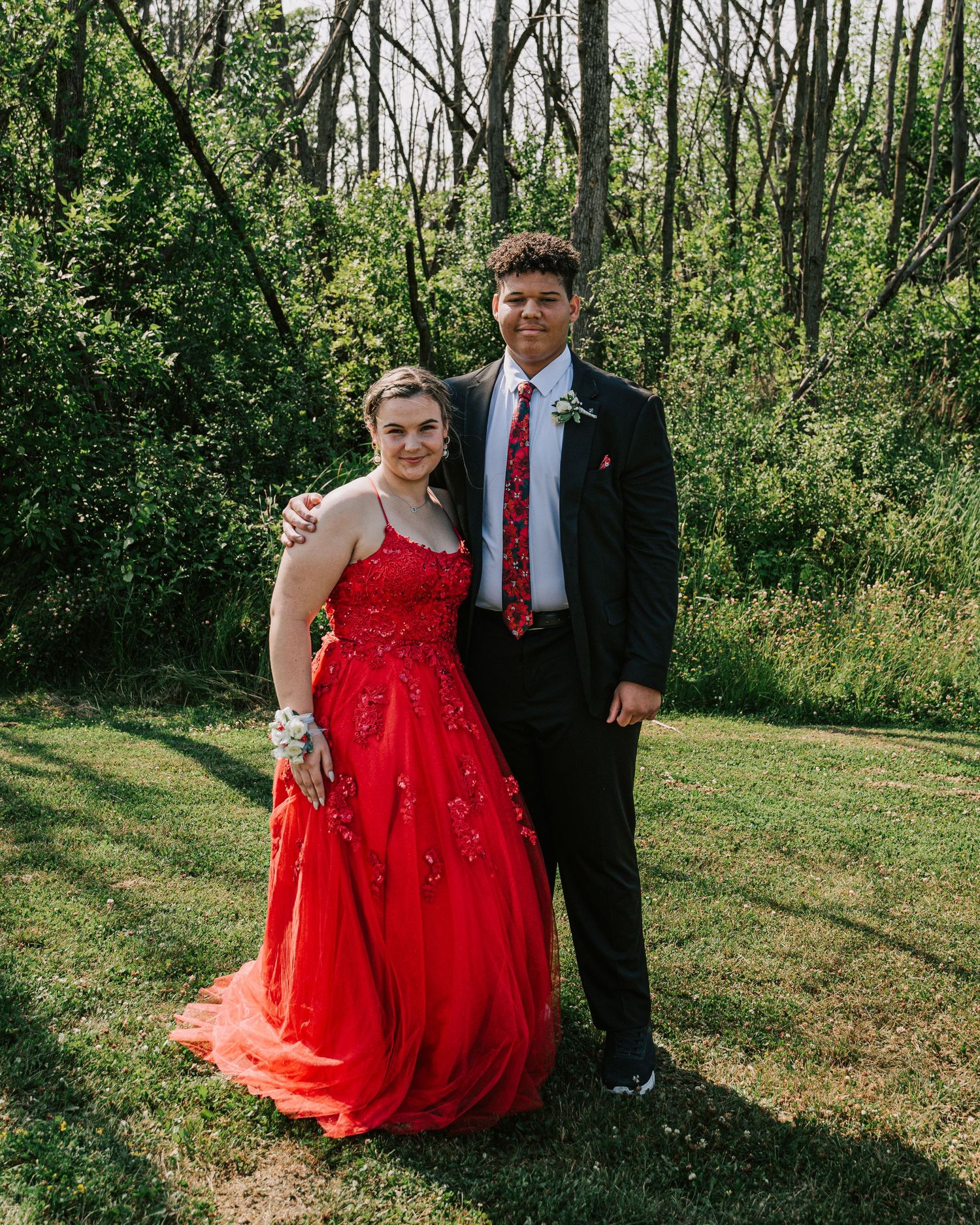 A couple poses together outdoors in formal attire with one wearing a sparkly red ball gown against a wooded backdrop.