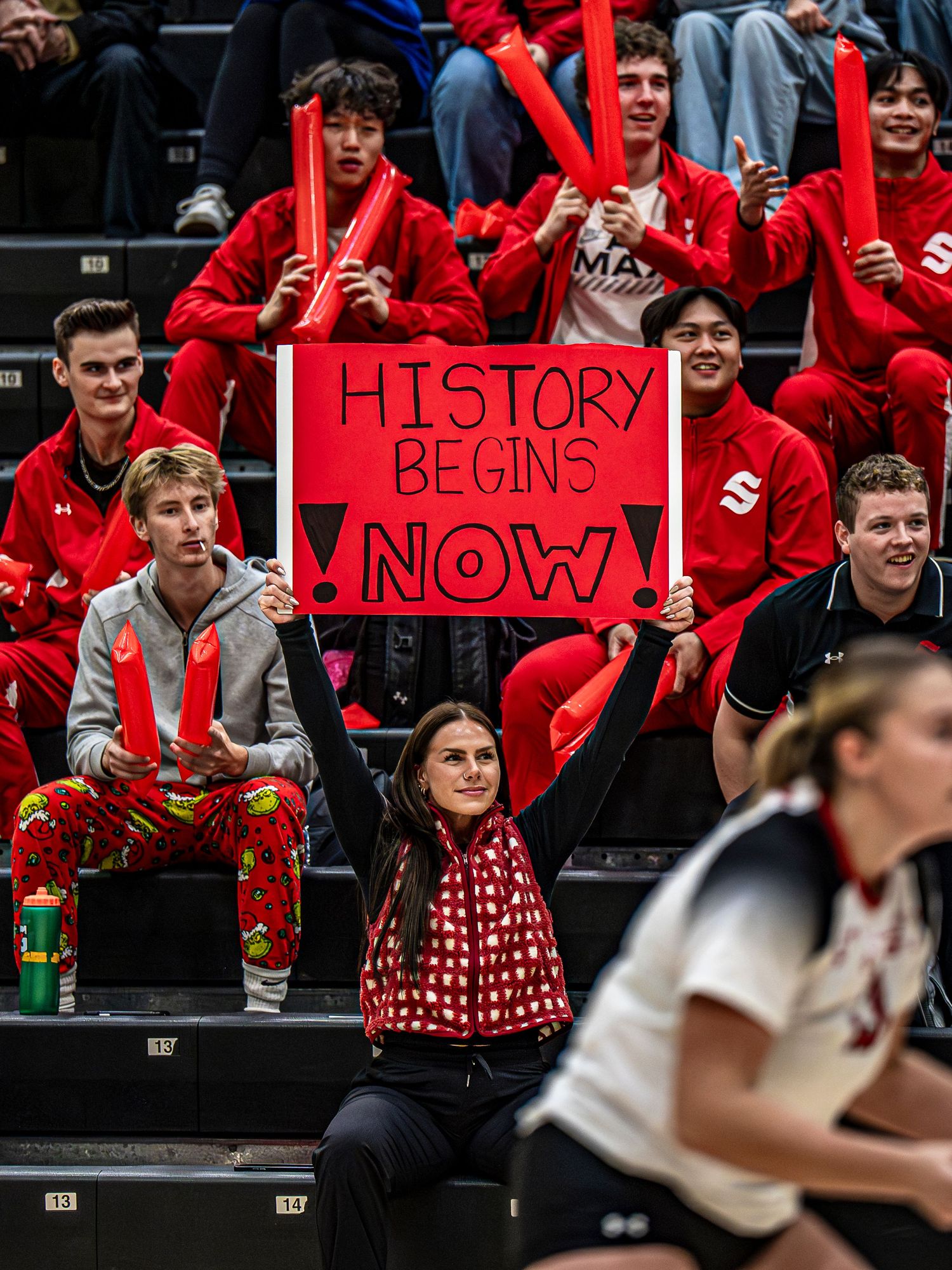 Crowd of fans in red clothing hold signs cheering at indoor sporting event.