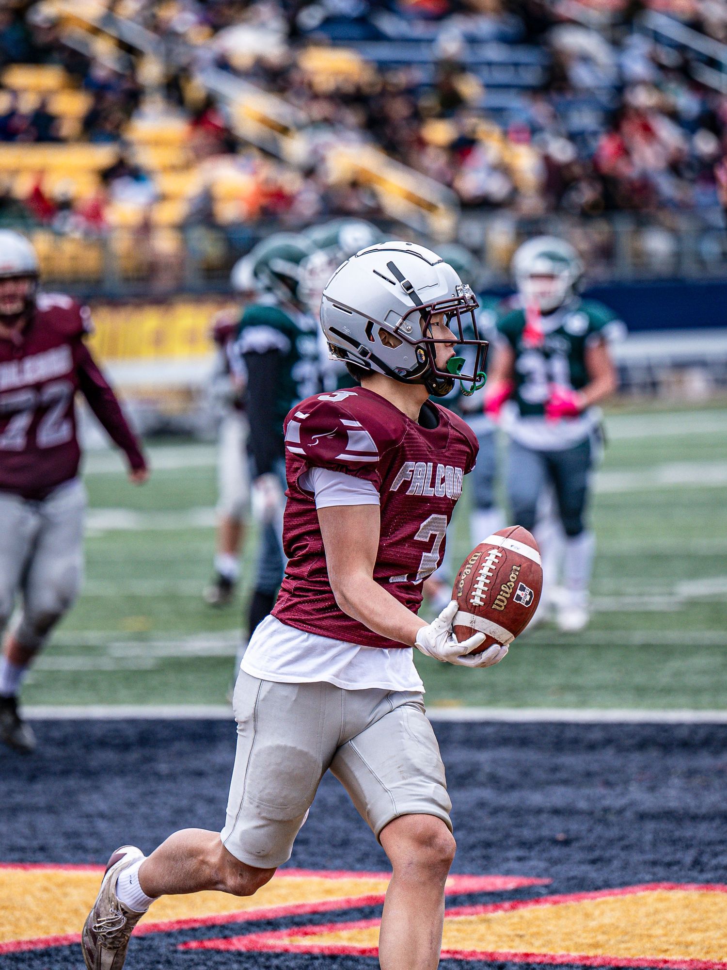 Football player in maroon uniform runs with ball during game on turf field.