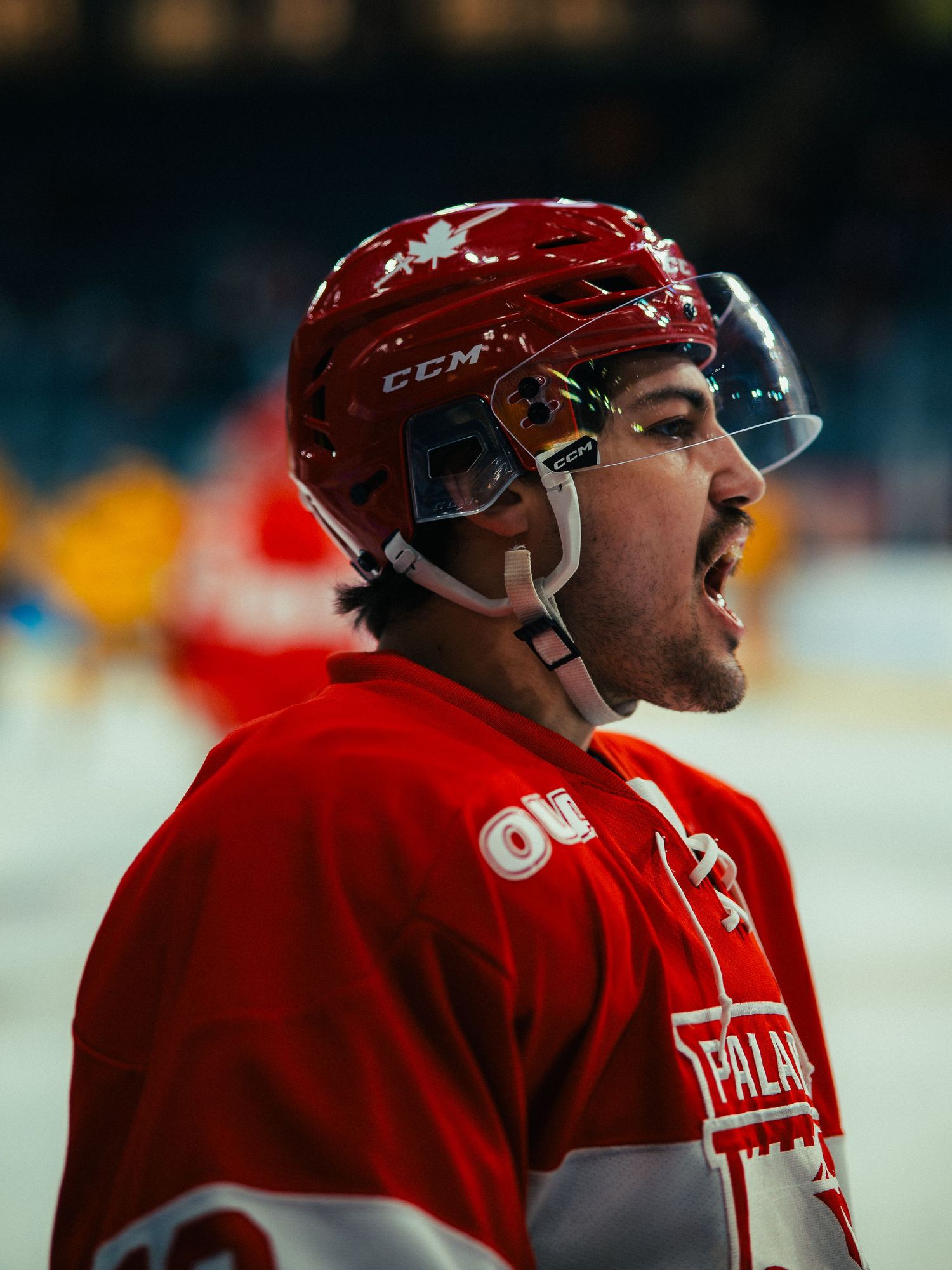Hockey player wearing a red CCM helmet and jersey yells during an intense moment on the ice.