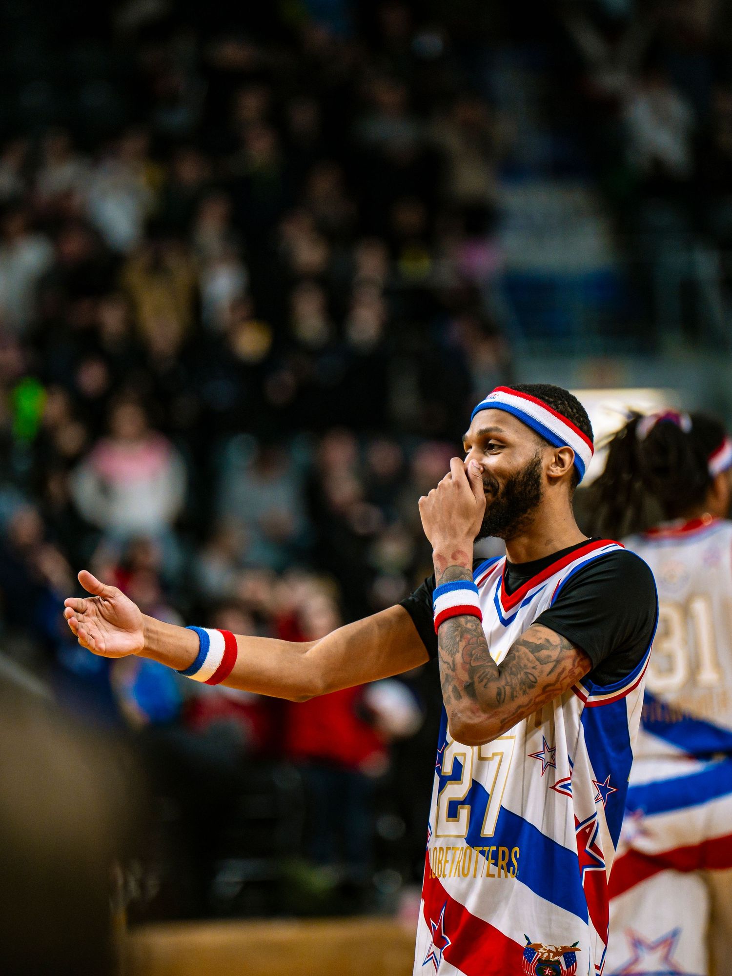 Basketball player in retro-style uniform with red and blue stripes gestures emotionally during game with crowd in background.