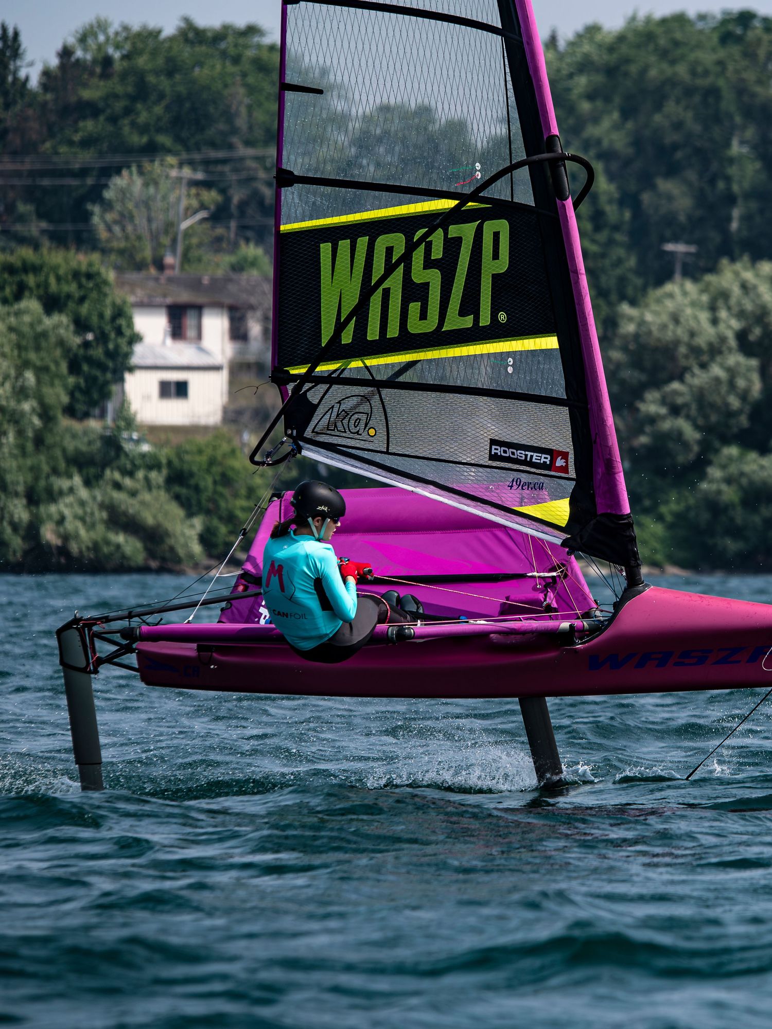 A bright pink hydrofoil sailboard with WASZP branding glides across blue waters near a wooded shoreline.