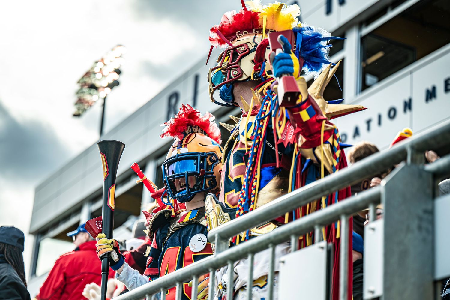 Crowd of sports fans in colorful attire cheer from stadium bleachers.