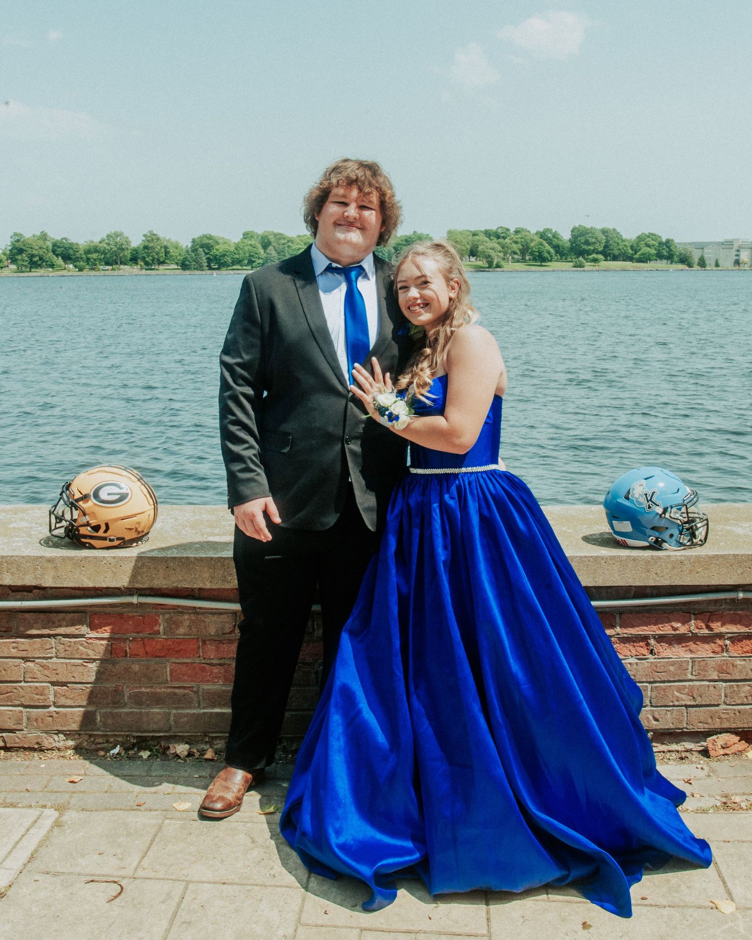 A couple in formal attire sits by a waterfront brick wall, with the person in a royal blue ballgown dress.