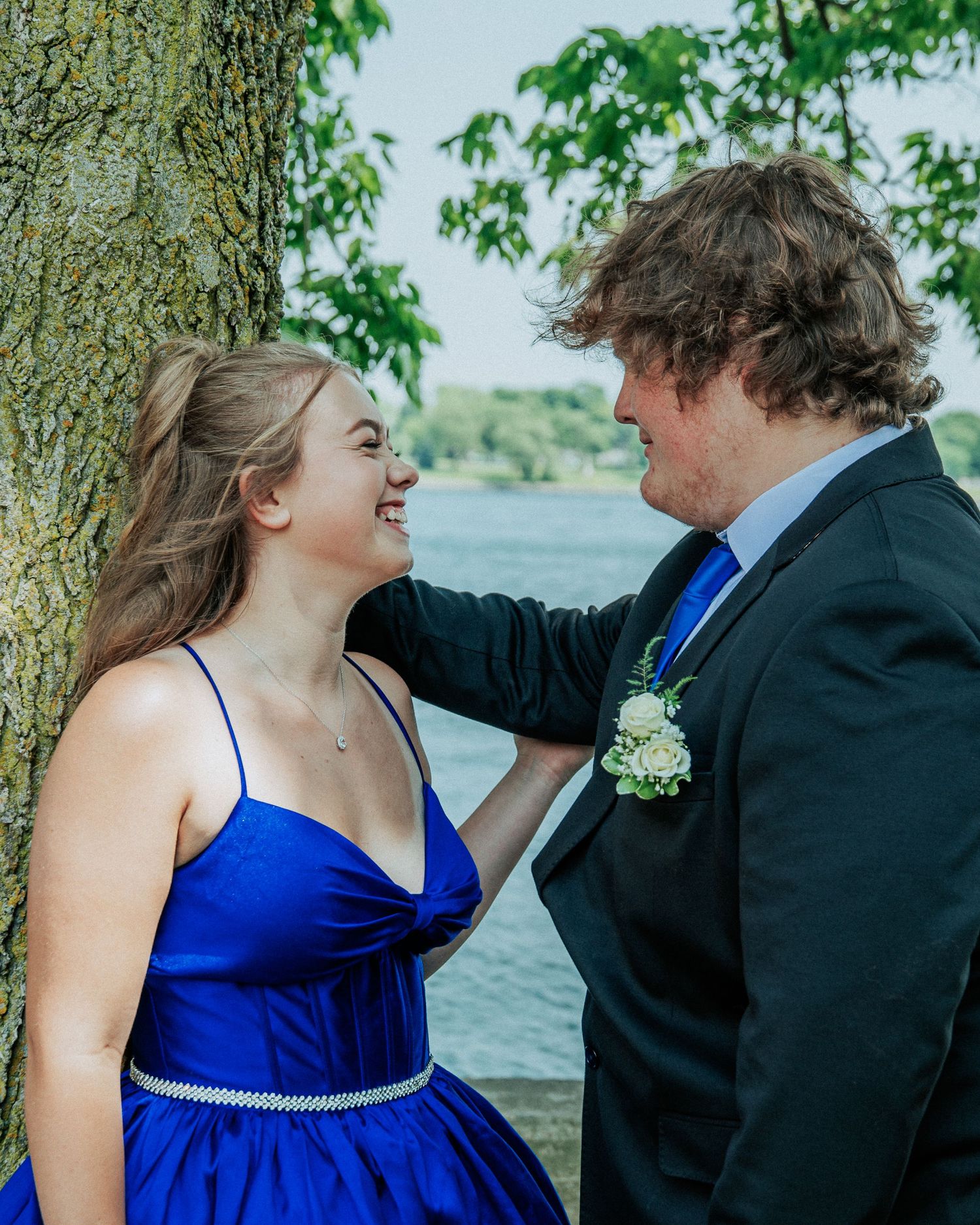 A couple in formal blue and black attire share candid moments by a lakeside with trees in the background.
