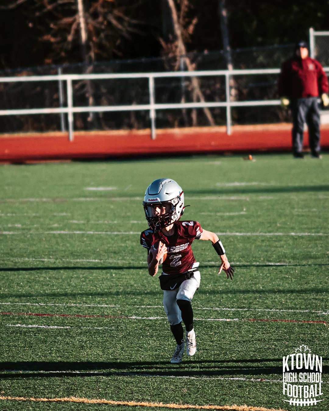 Youth football player in white and maroon uniforms crouches on artificial turf field during daytime practice.