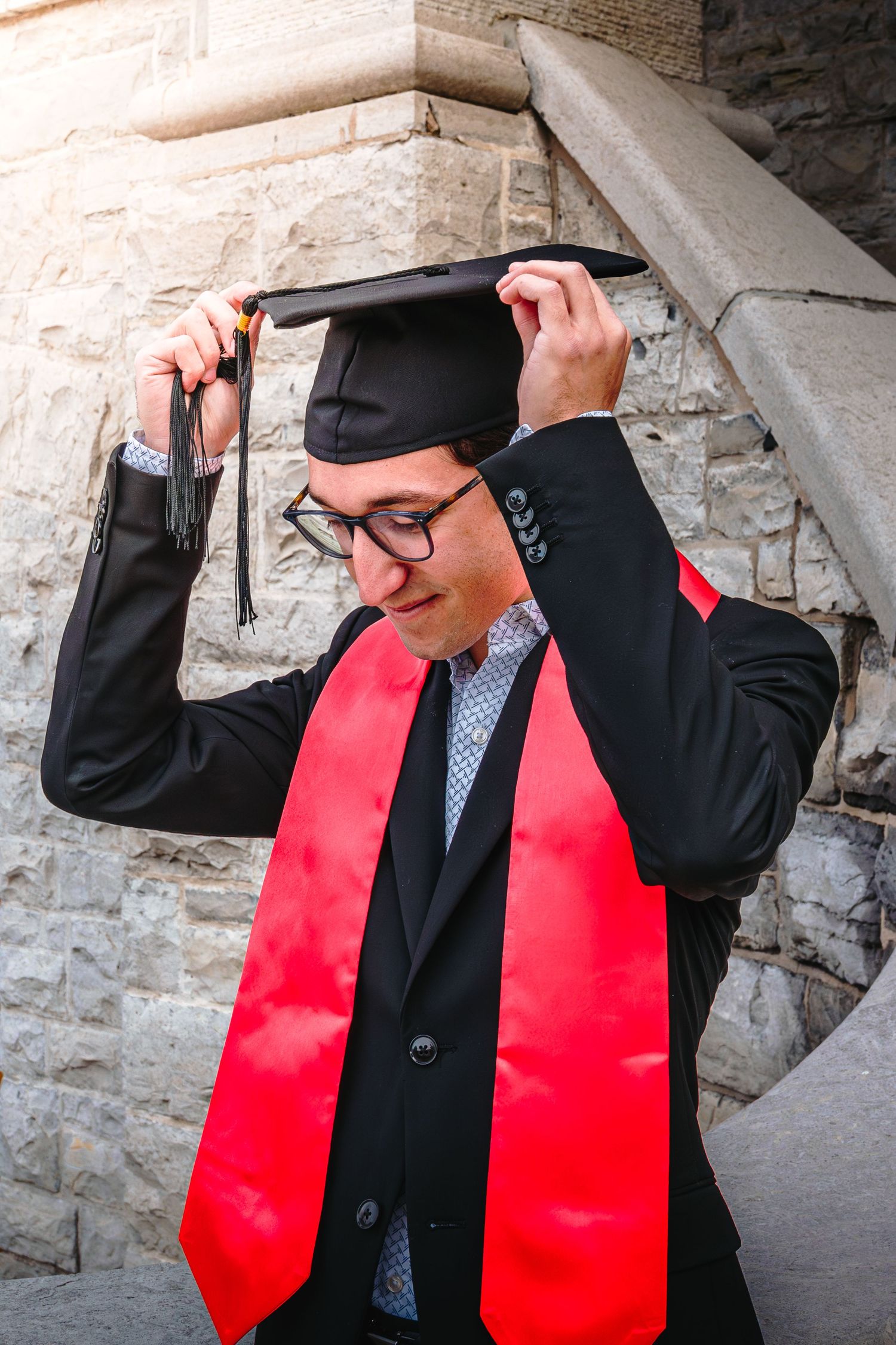 A graduate in black suit and red stole adjusts his graduation cap against a stone wall background.