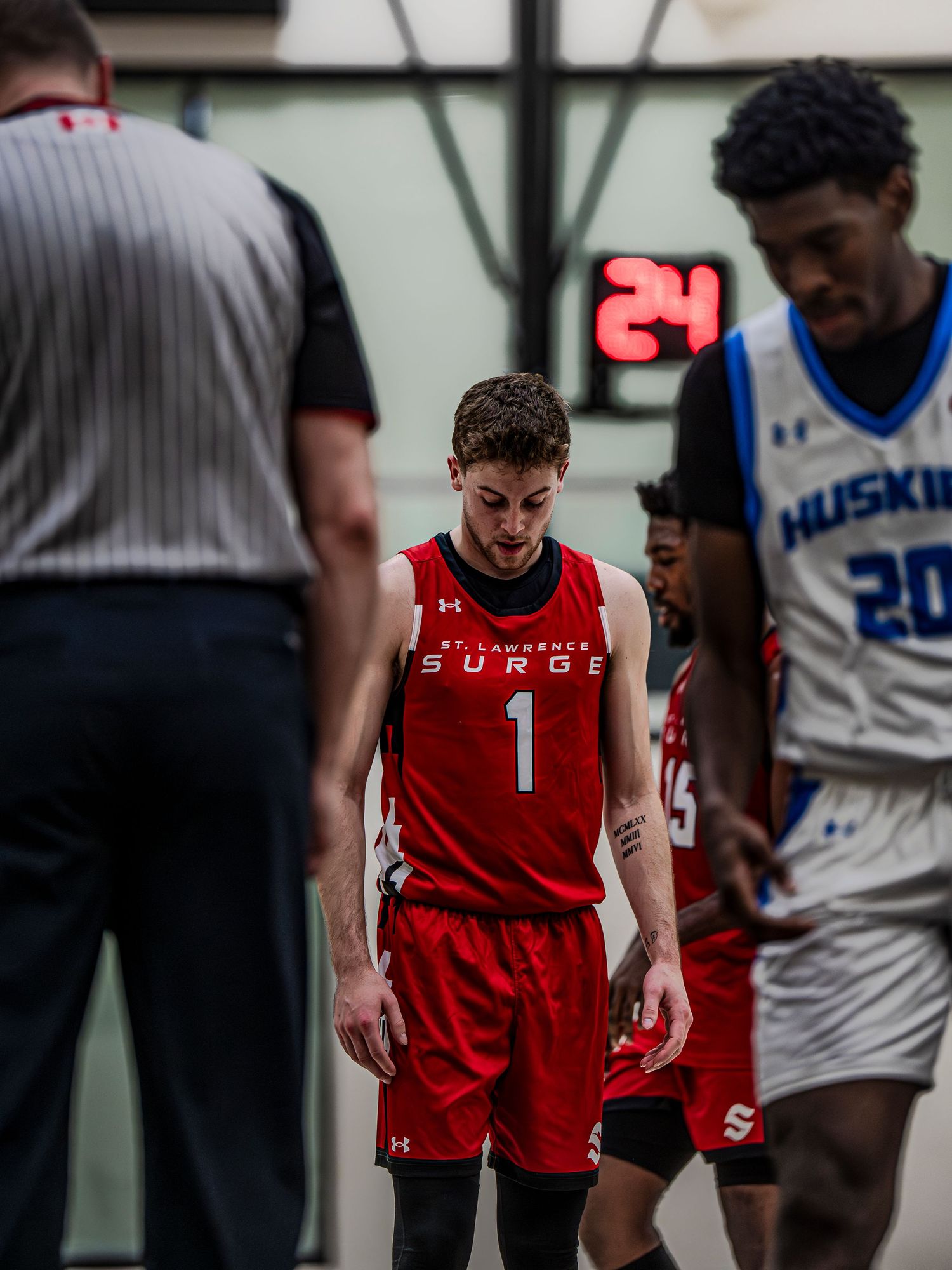 Basketball player in red jersey stands between referee and opponent during game stoppage.