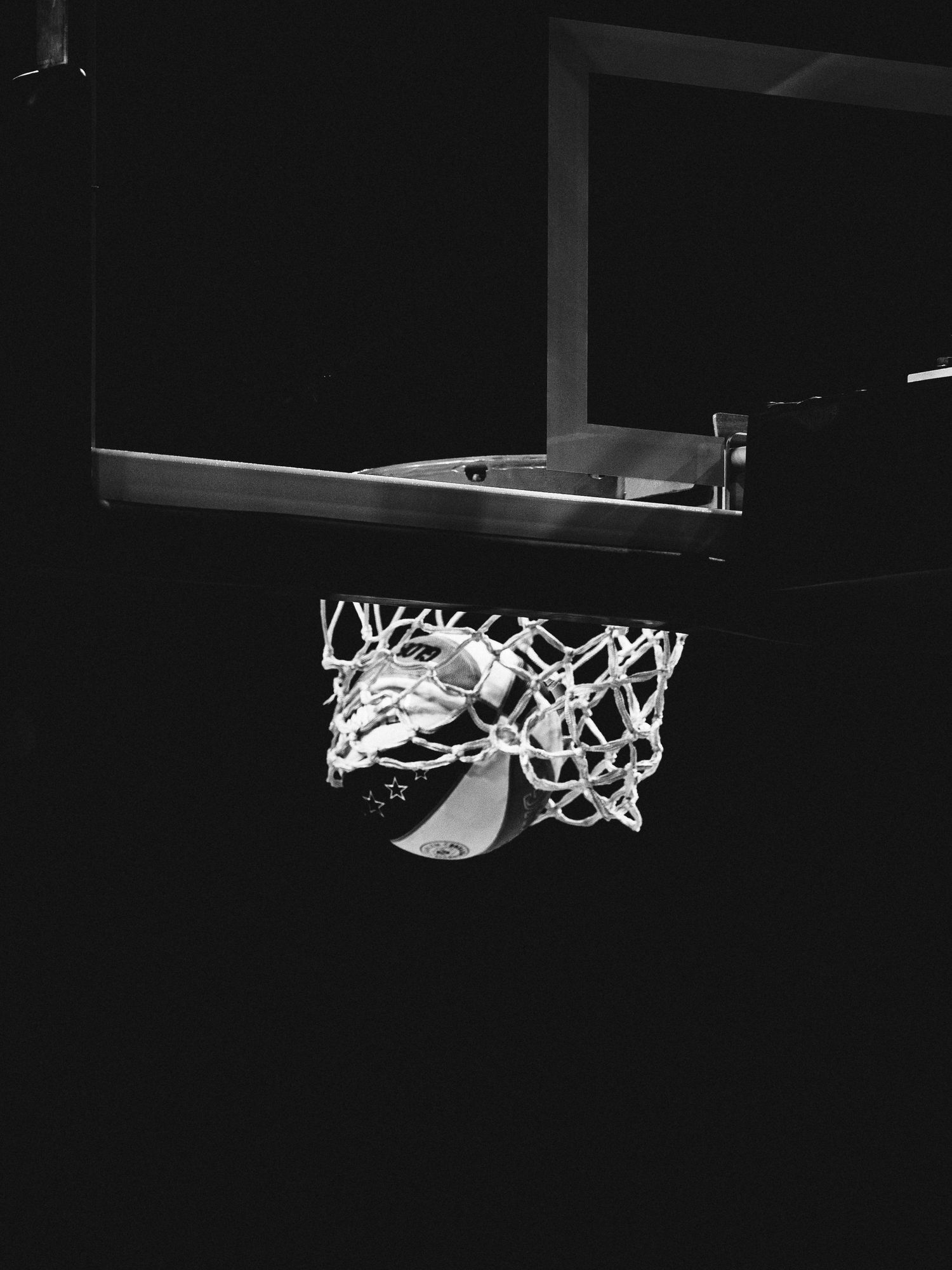 Black and white photo of a basketball passing through a net on a hoop against a dark background.