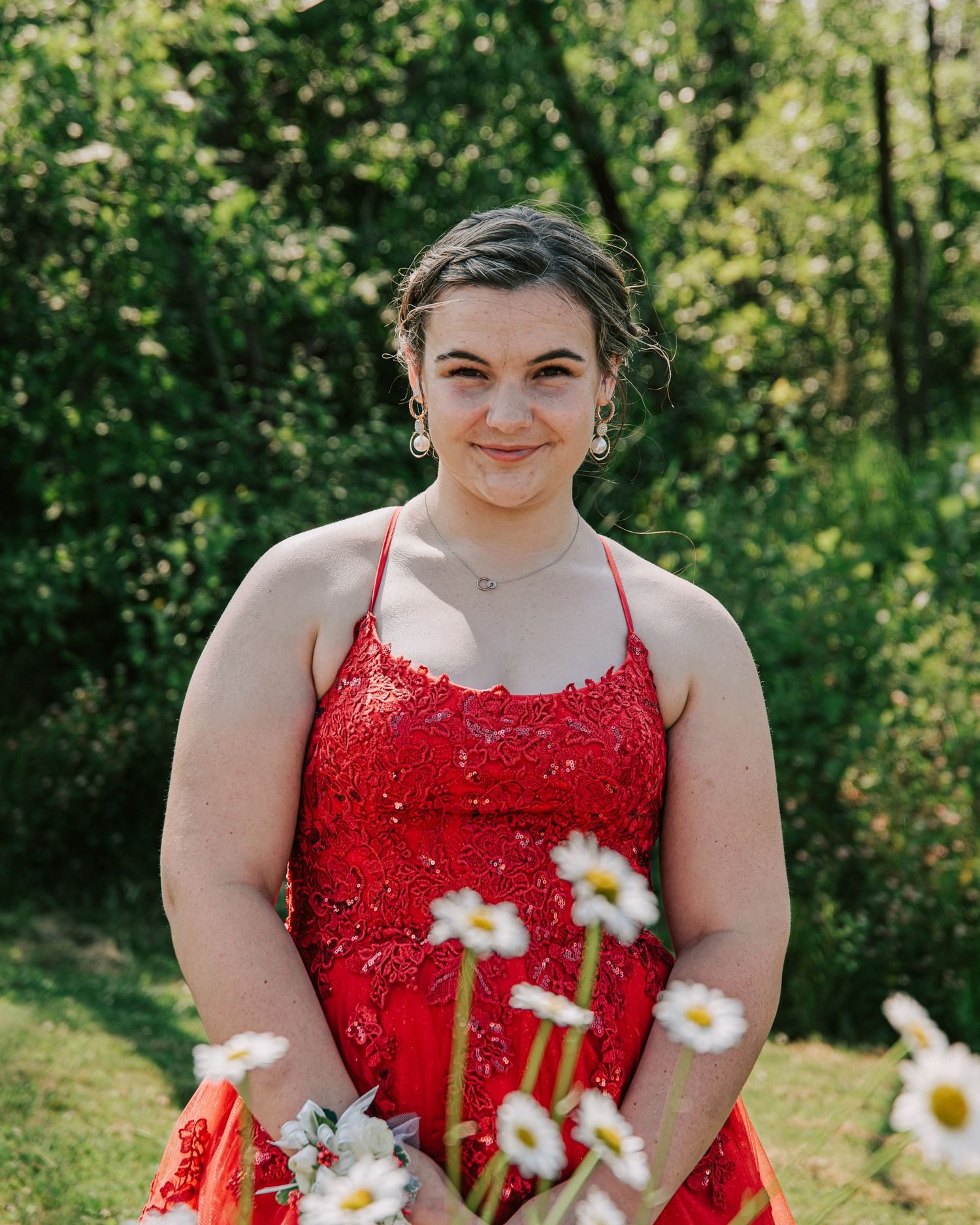 Person in a sparkly red formal dress poses outdoors with white daisies in a sunny garden setting.