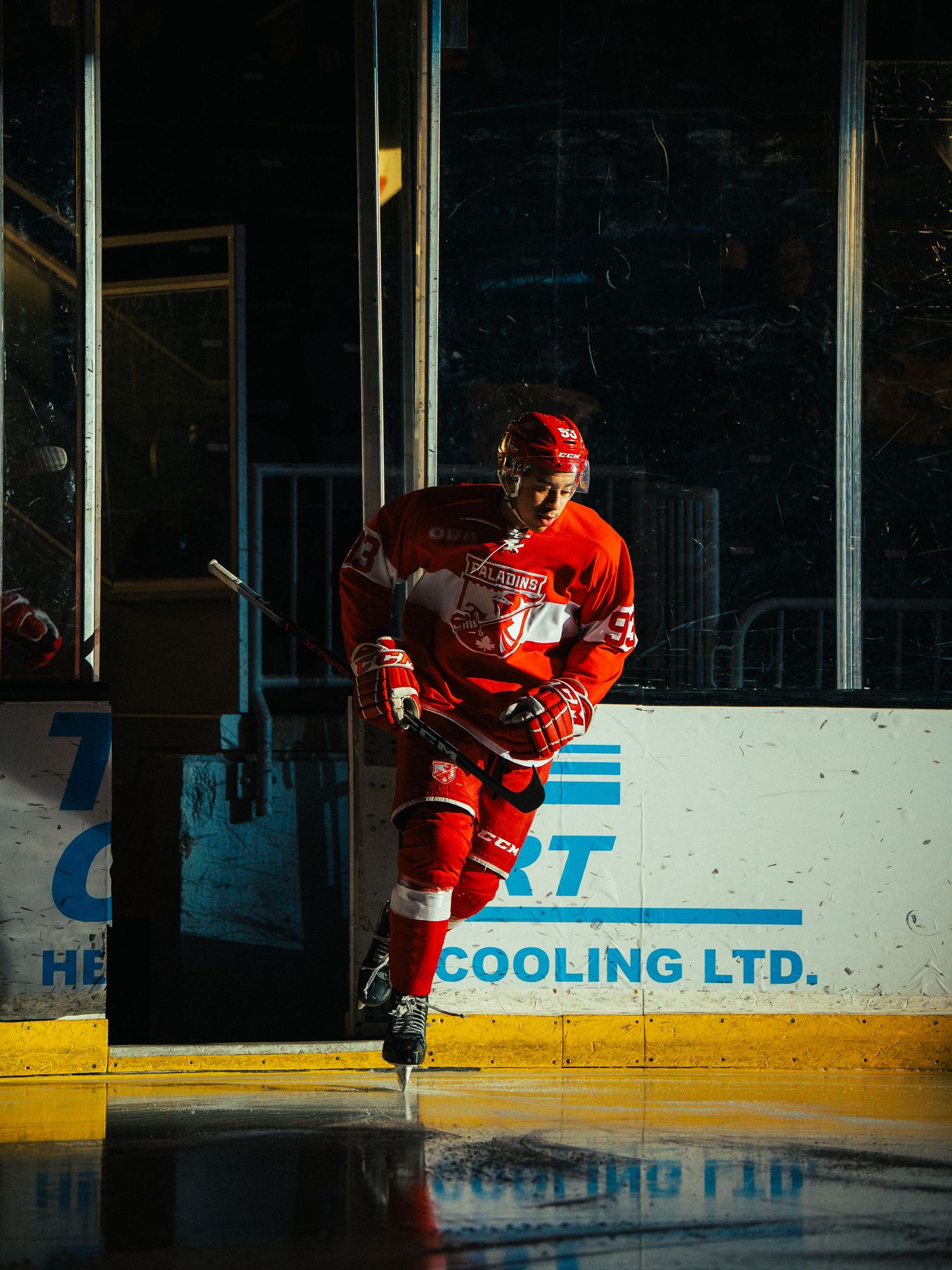 Hockey player in red uniform skating on ice rink near boards with advertising signage visible.