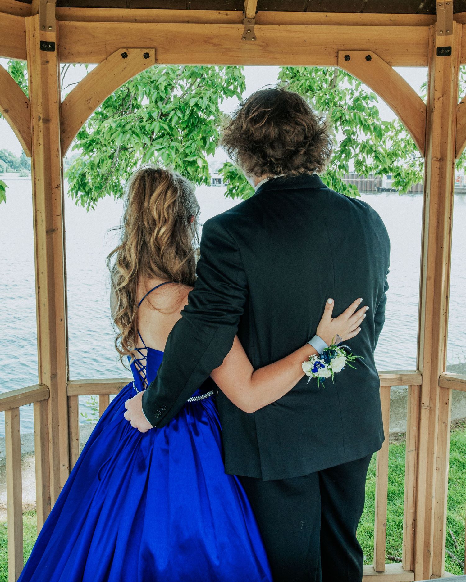 A couple in formal attire embrace while standing in a wooden gazebo overlooking a lake, with lush greenery in the background.