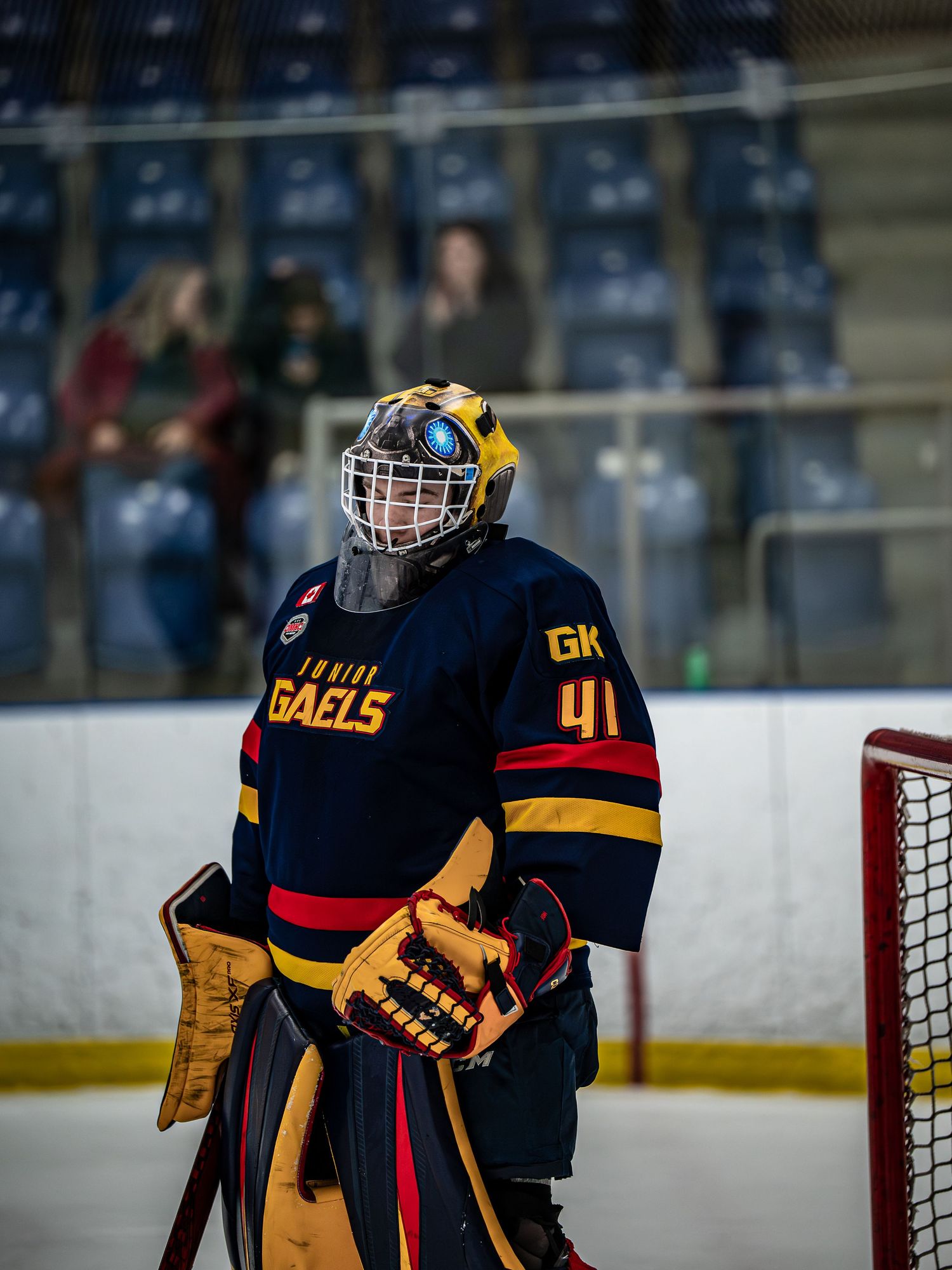 Hockey goalie in navy blue and yellow uniform stands ready at the net during an indoor ice hockey game.