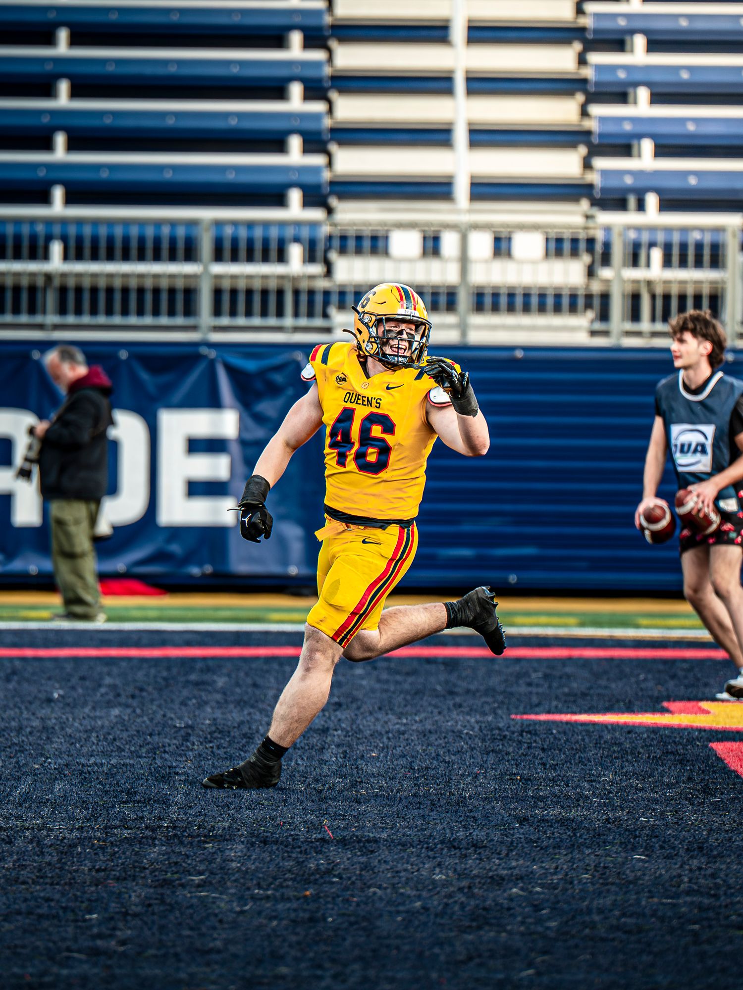 Football player in yellow jersey dives across black turf field during game action.
