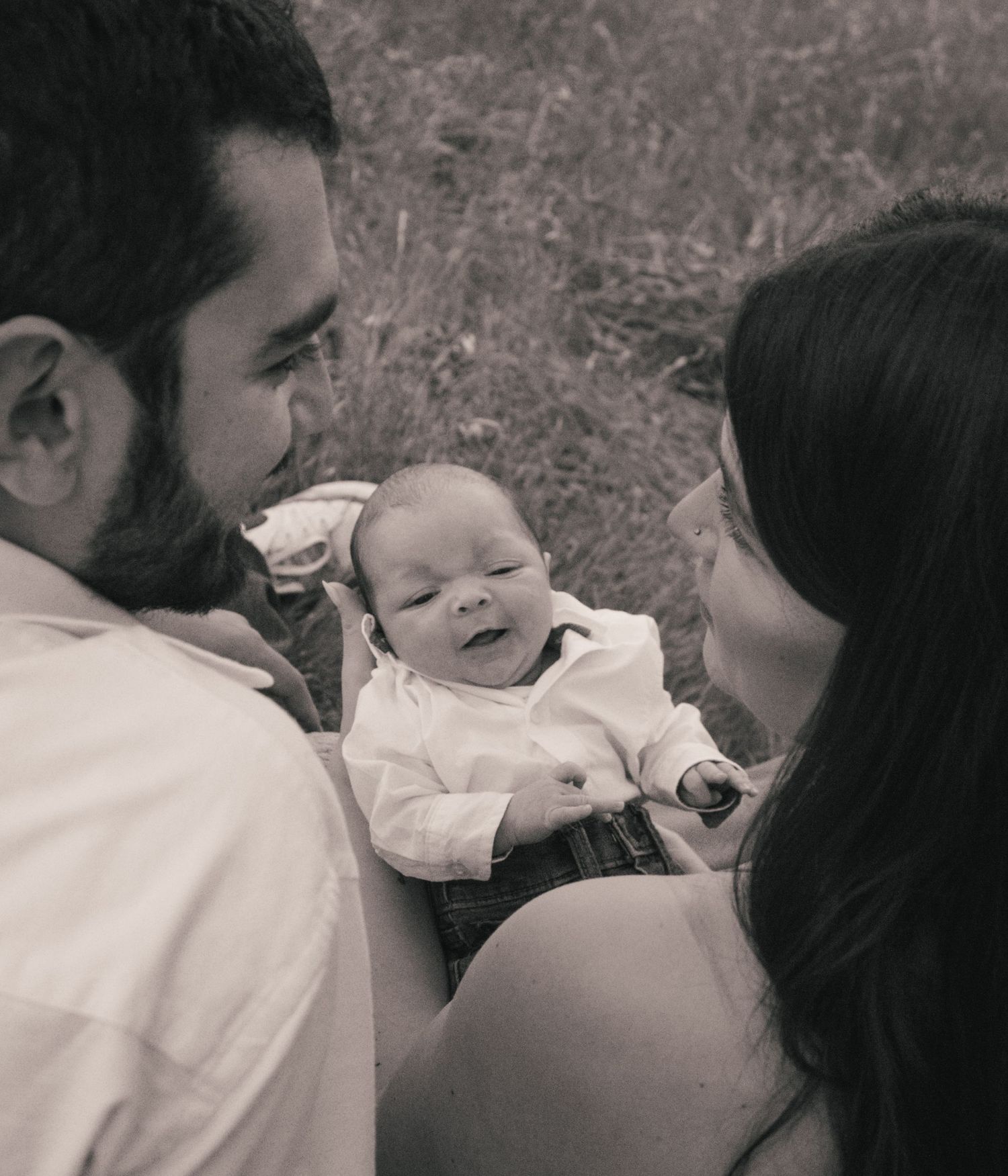 Black and white photo of young parents smiling at their newborn baby outdoors in a field.
