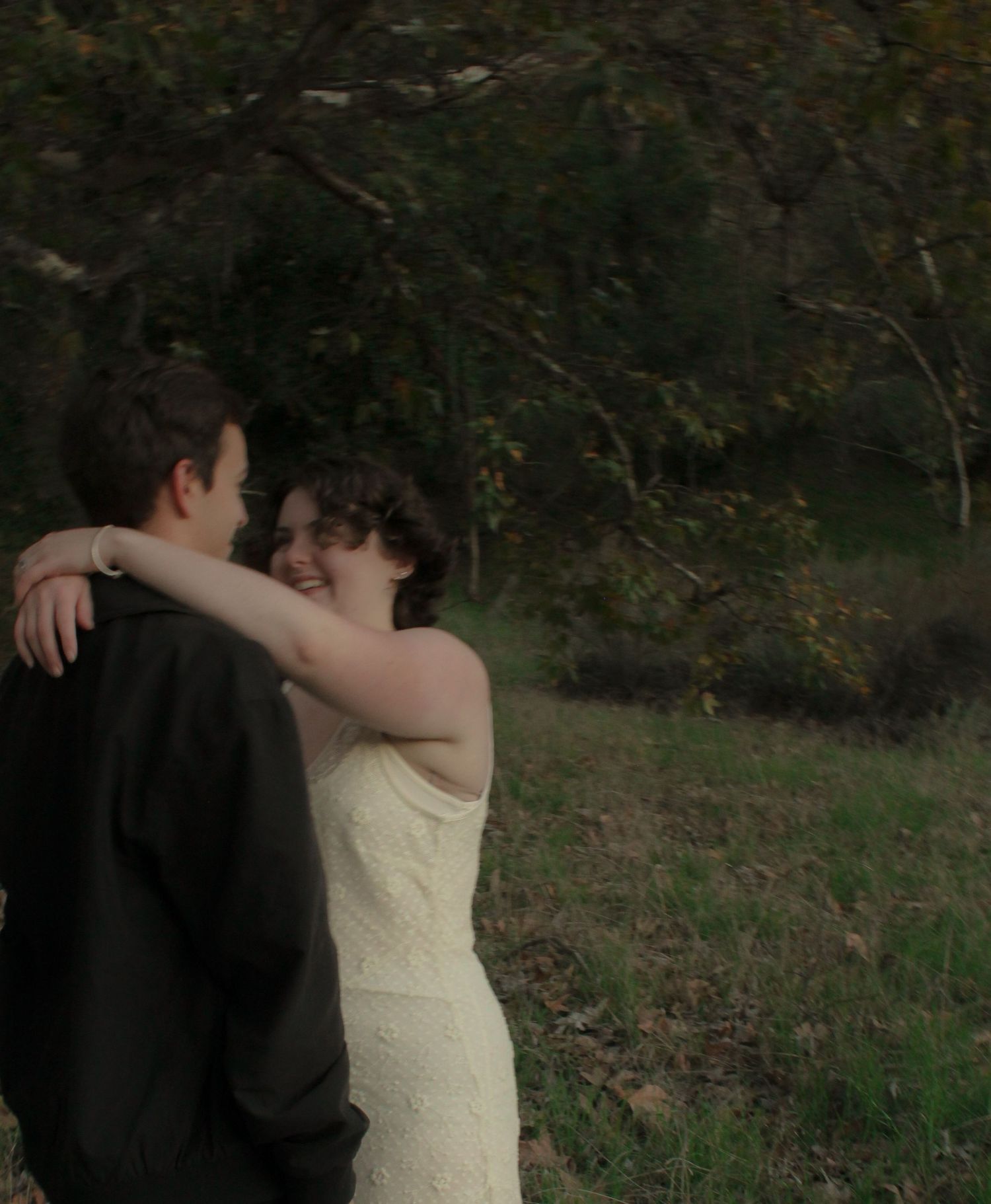 A couple in formal attire embraces and dances together in an outdoor setting with trees in the background.