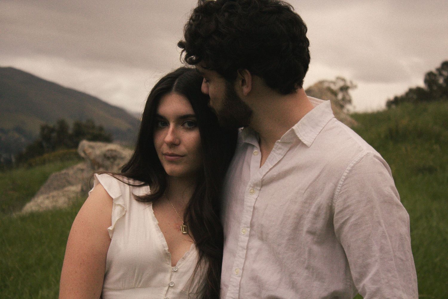 A couple embraces in a romantic outdoor setting with rolling hills and natural scenery in the background.