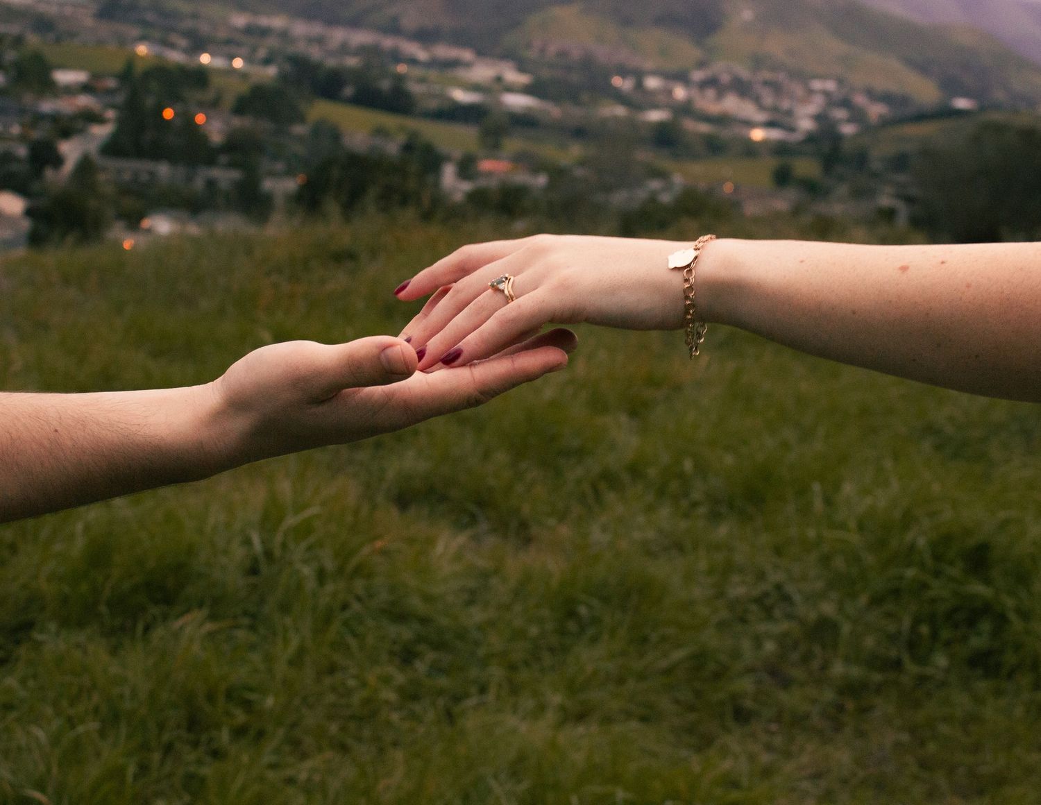 Two hands reaching towards each other in a grassy field with mountains in the background at sunset.