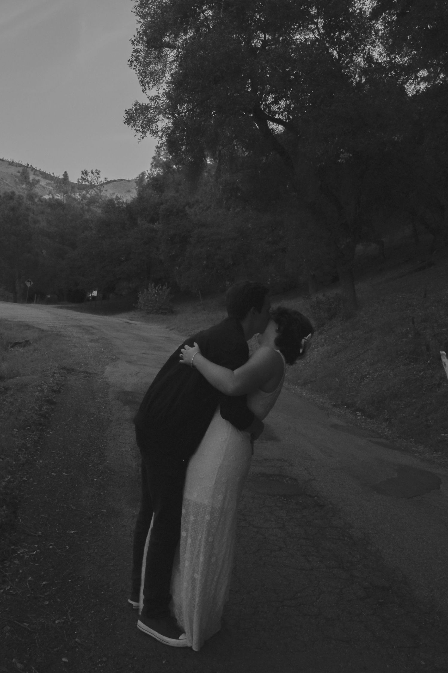 A romantic black and white photograph of two people embracing on a winding rural road surrounded by trees.