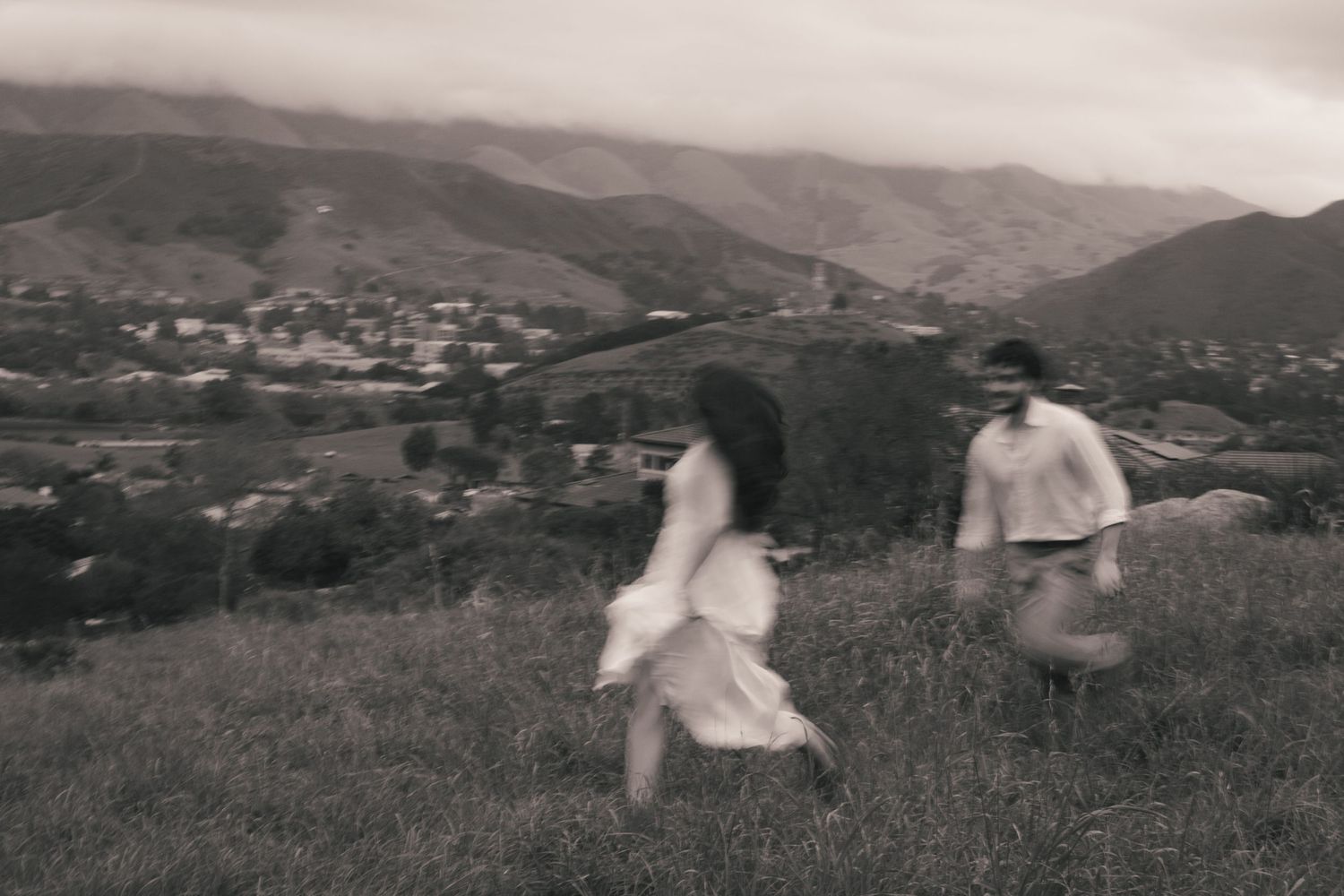 A blurry black and white vintage photograph shows people running across a grassy hillside with mountains in the distance.